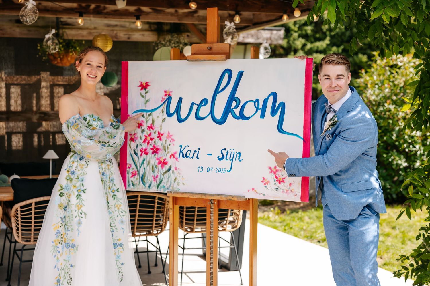 Bride and groom smile beside a colorful welcome sign reading Welkom Kari-Stijn at their wedding.