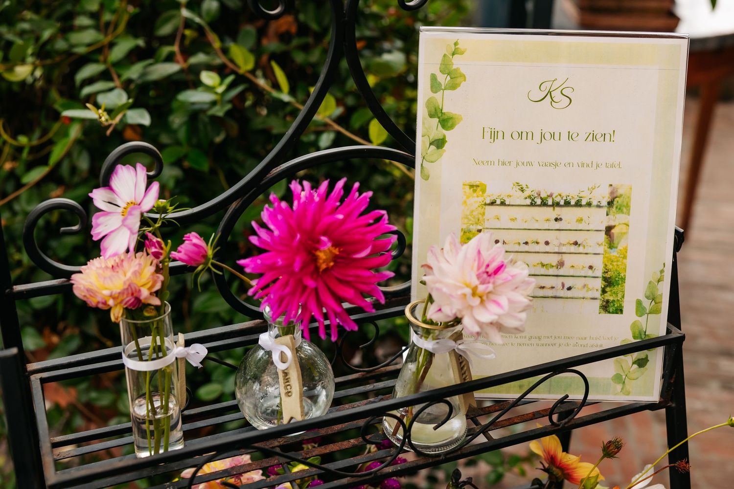 Pink dahlias and wildflowers in glass bud vases beside wedding sign on iron shelf.