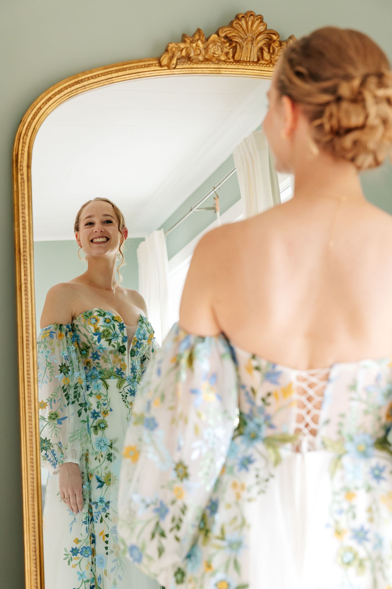 Bride in floral off-shoulder gown smiling at her reflection in an ornate gold mirror.