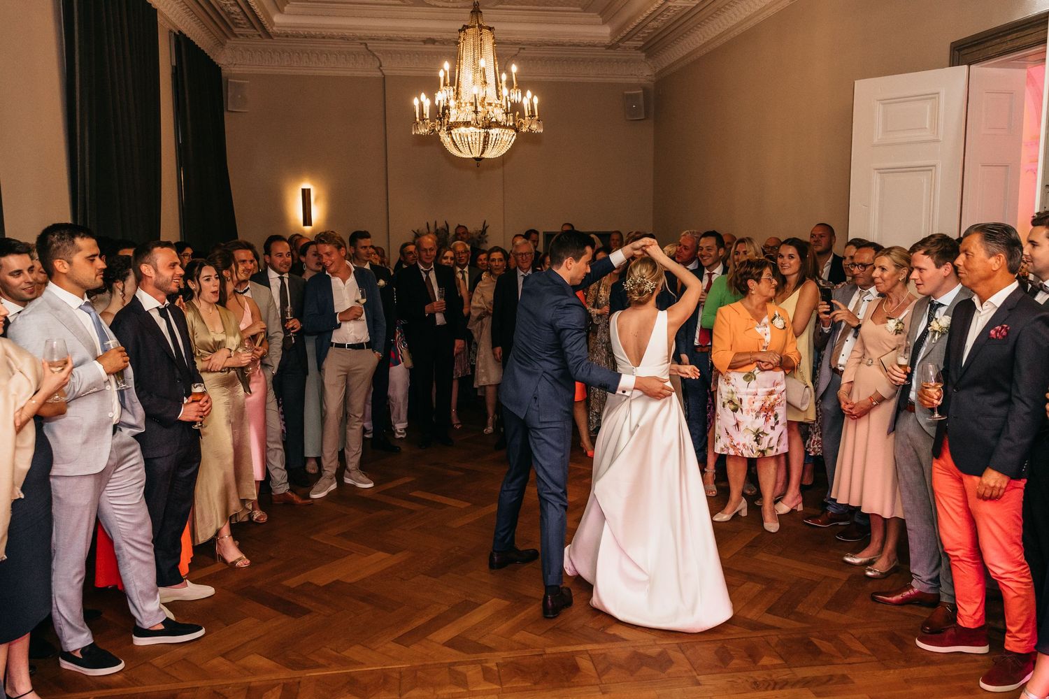 Bride and groom share first dance surrounded by wedding guests in elegant ballroom with chandelier.