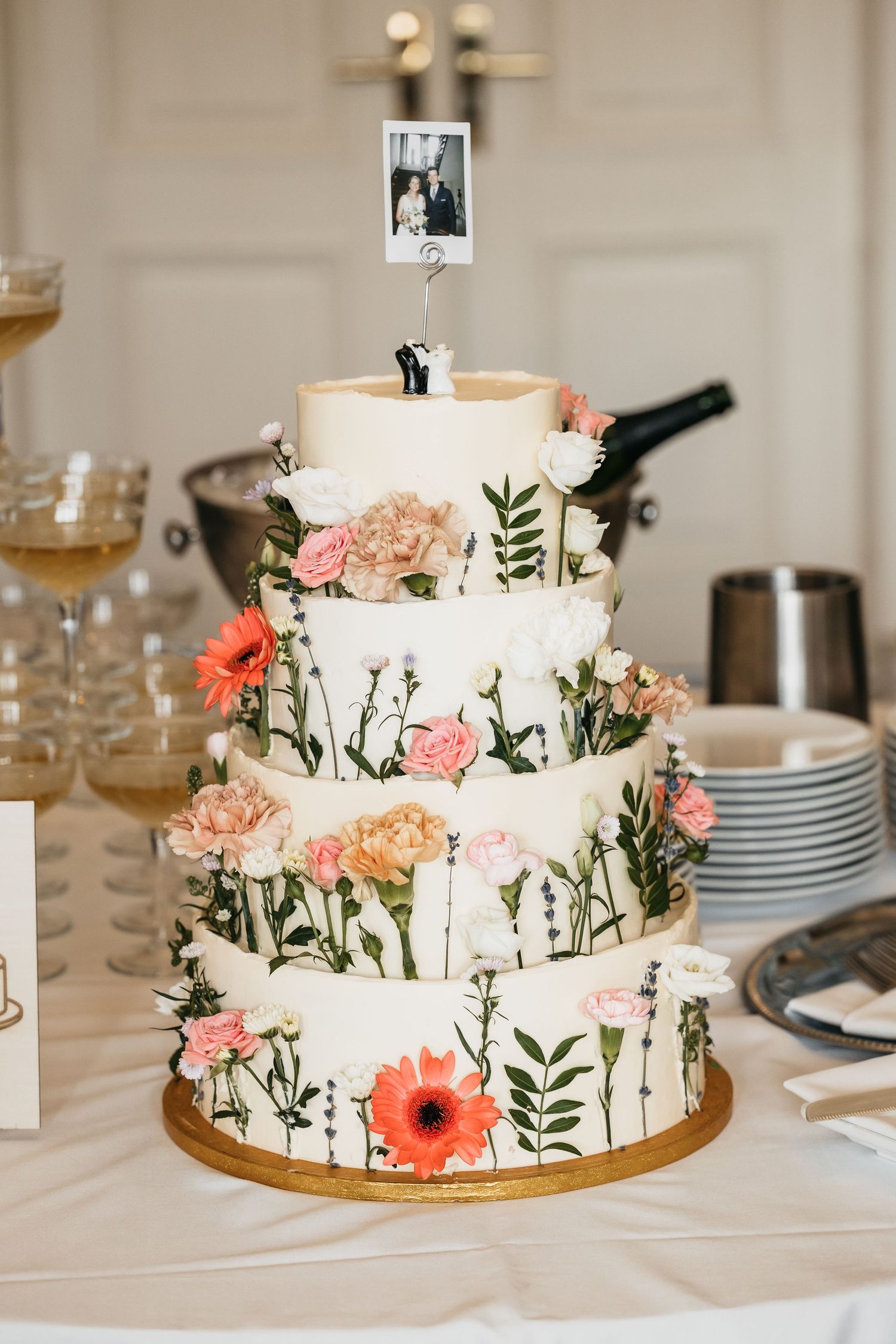 Four-tier white wedding cake decorated with colorful wildflowers and a Polaroid photo topper.