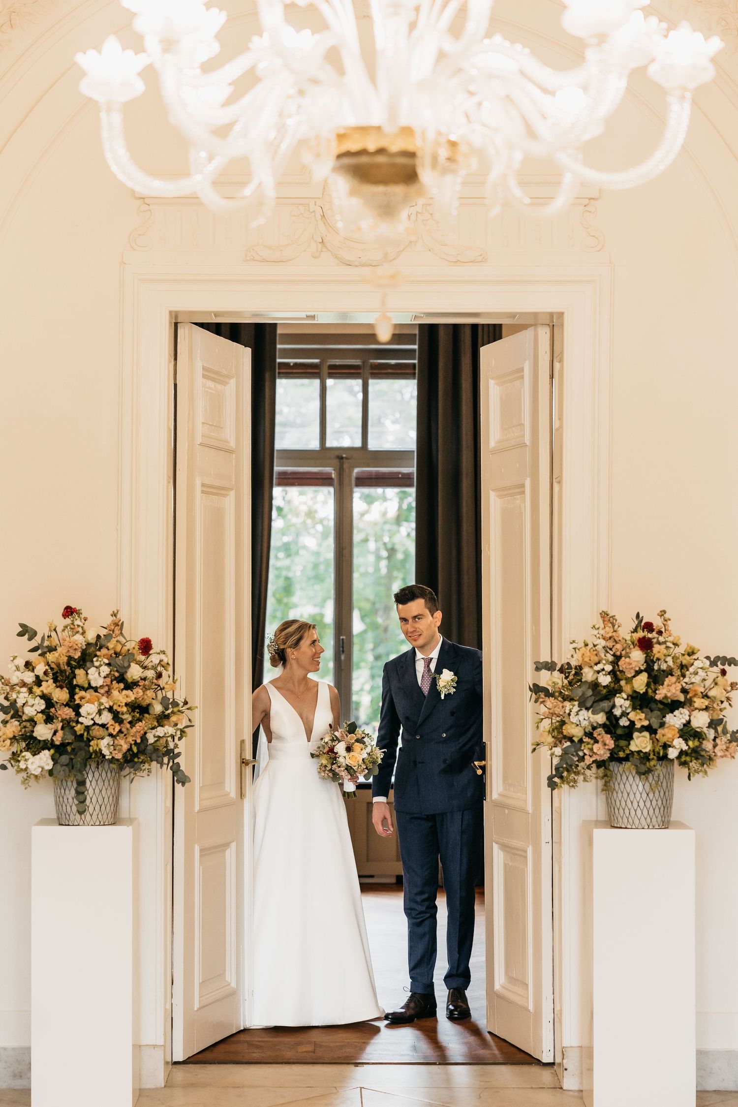 Bride in white gown and groom in navy suit stand in ornate doorway flanked by floral arrangements.