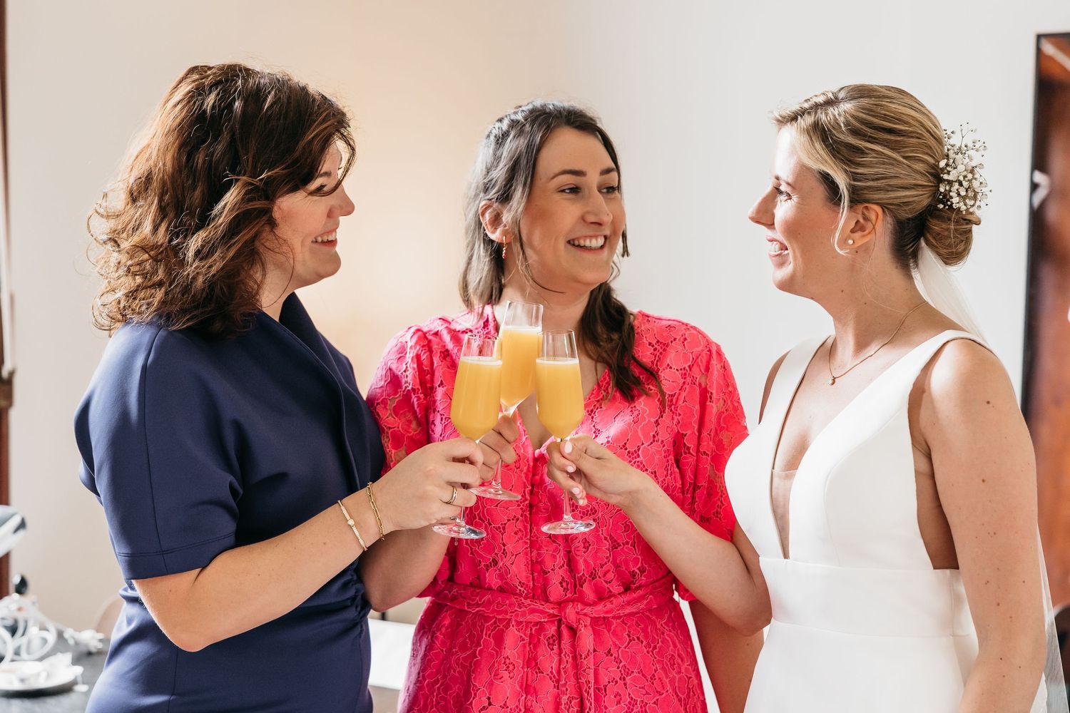 Bride and two women in navy and pink outfits cheerfully toasting with mimosa glasses.