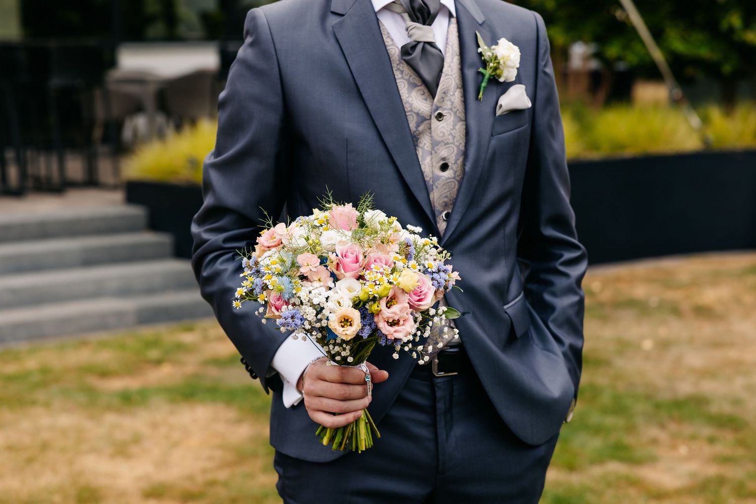Groom in navy suit holds colorful bridal bouquet with pink and white flowers outdoors.