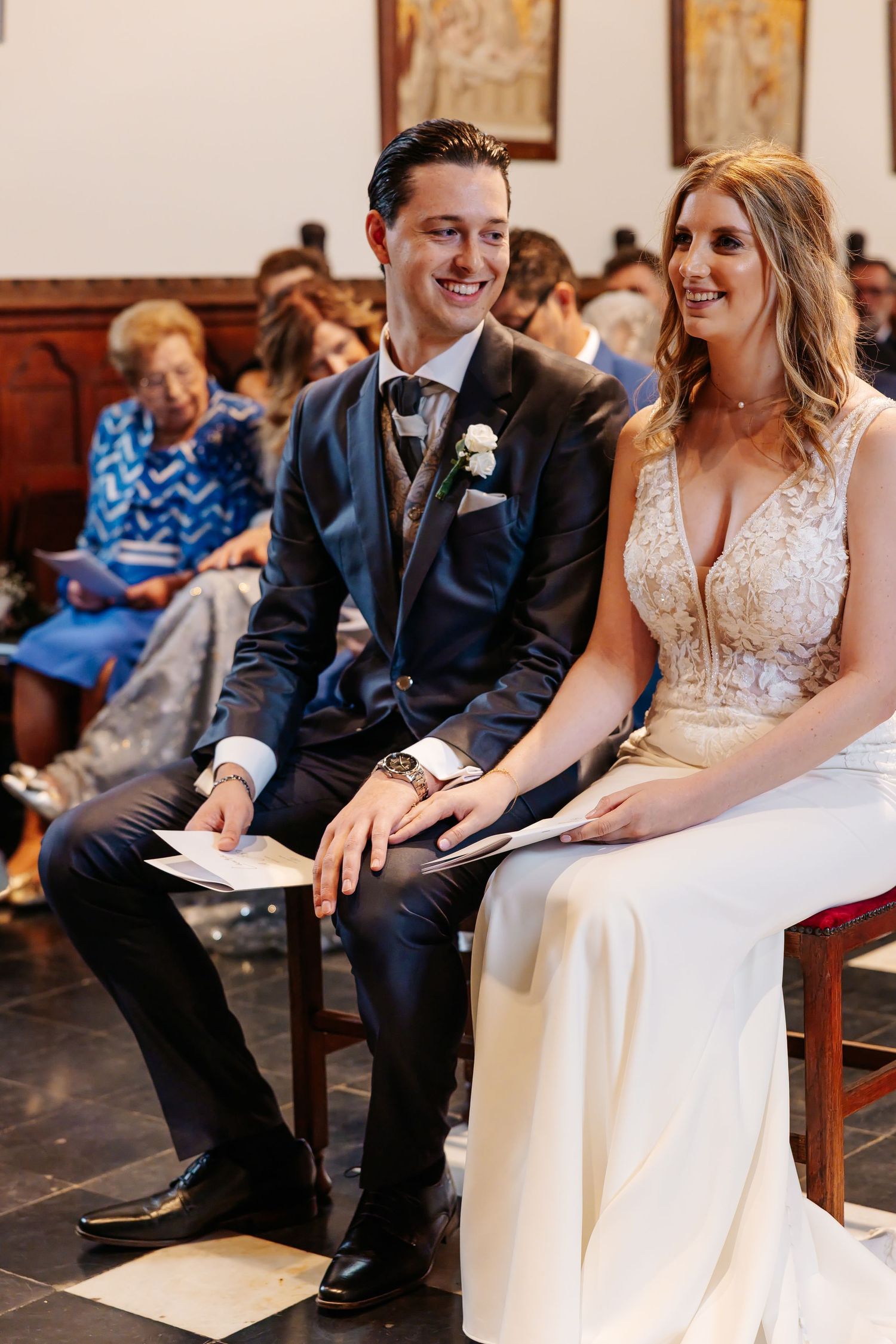 Bride and groom sit together smiling during church wedding ceremony holding programs.