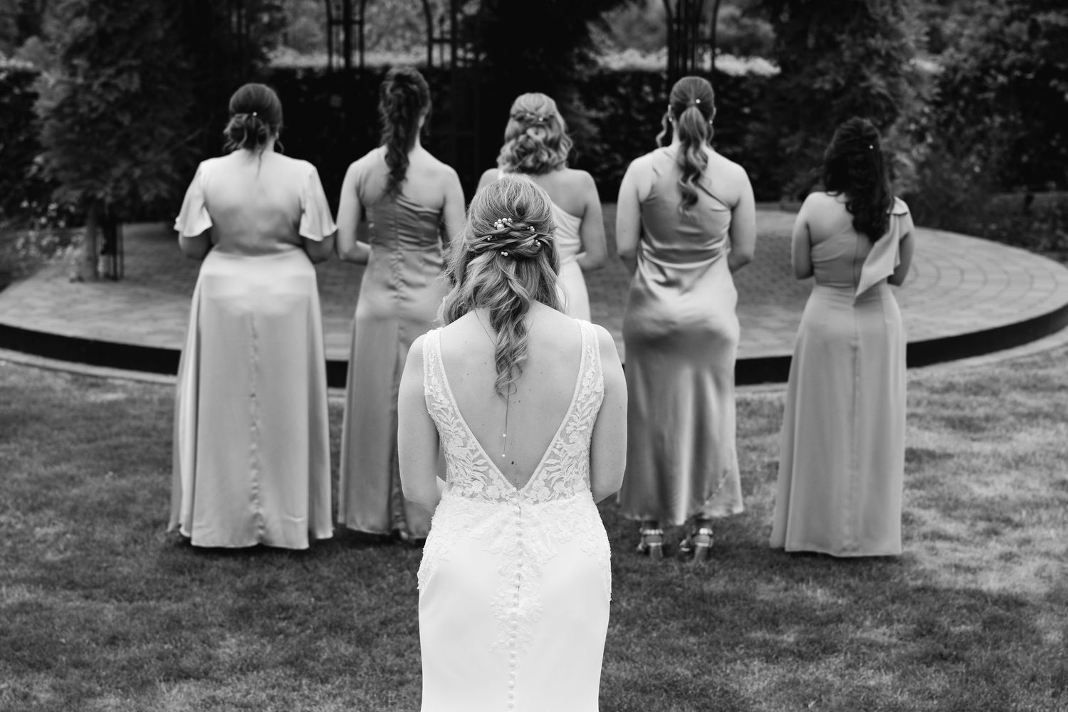 Black and white photo of bride with lace back gown facing bridesmaids near garden gazebo.