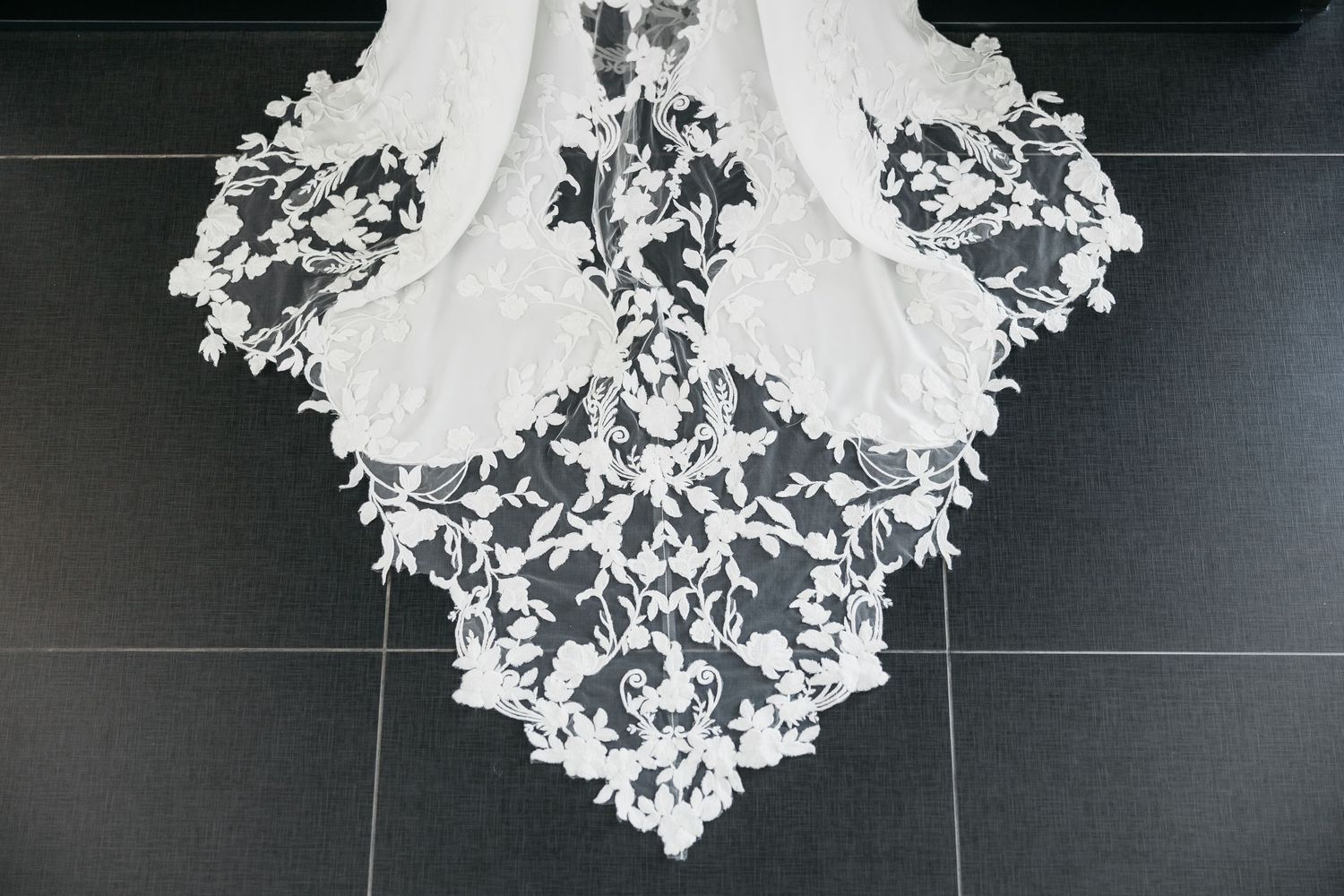 Detailed white embroidered lace bridal gown train displayed on black tile floor.