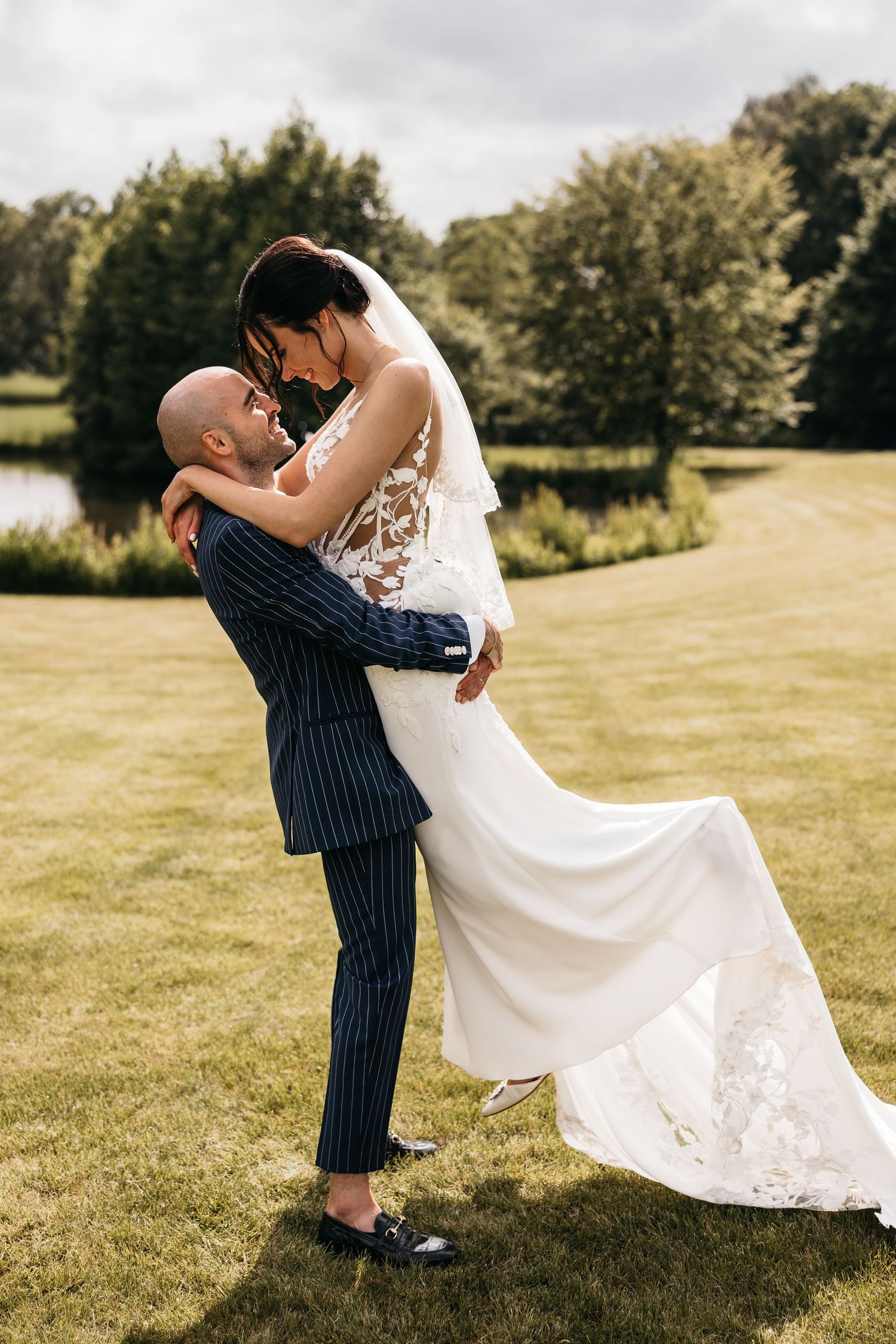 Groom in navy pinstripe suit lifting bride in lace wedding dress outdoors on green lawn.