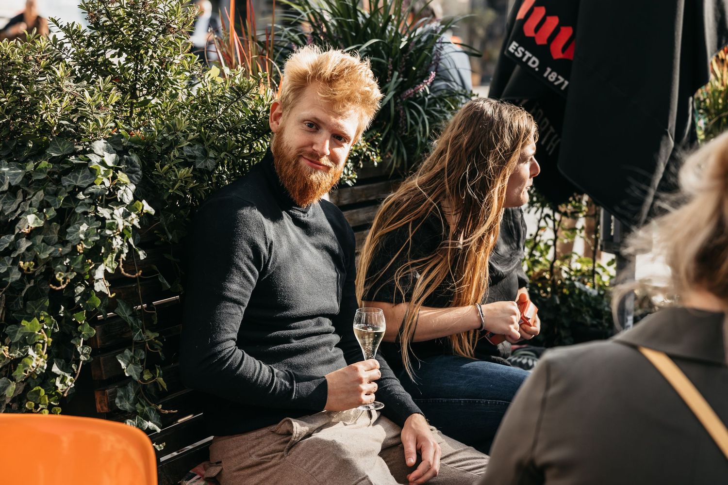 Red-haired bearded man holding champagne glass relaxing outdoors with friends at wedding reception.