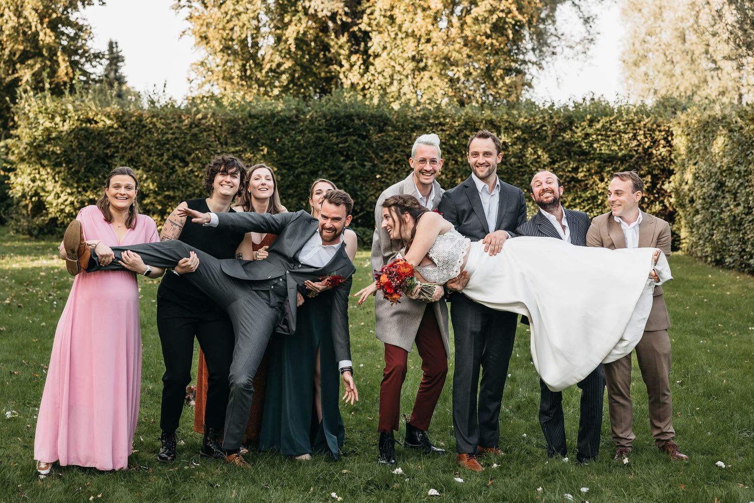 Groomsmen lifting bride and groom playfully outdoors at wedding celebration with bridal party on lush green lawn.
