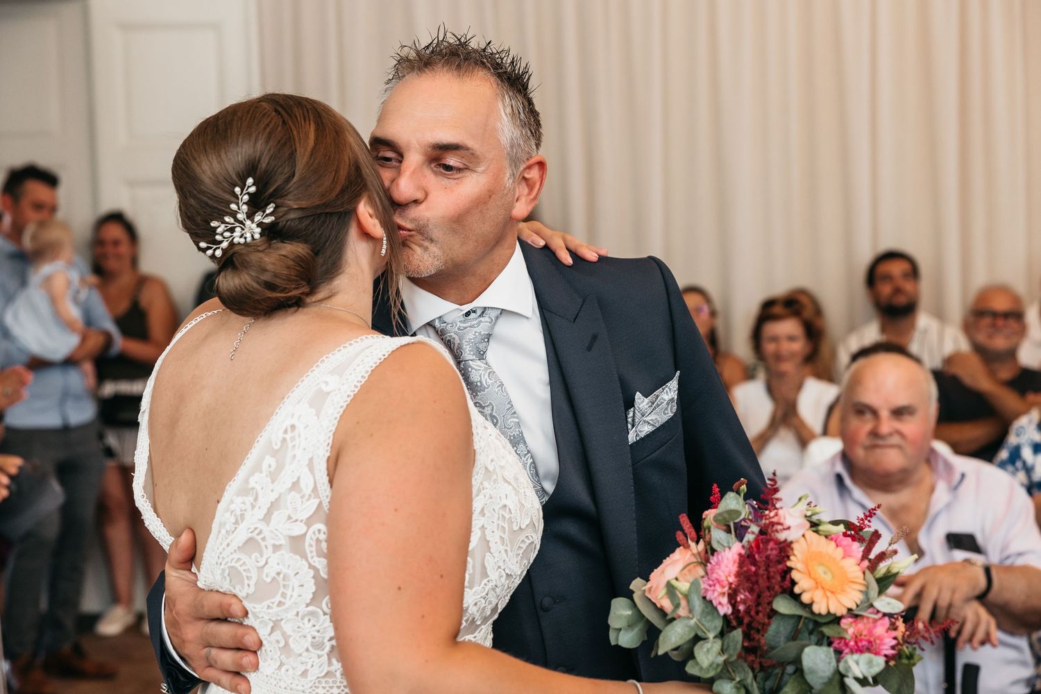 Groom in navy suit sharing a kiss with bride holding a colorful bouquet during wedding ceremony.