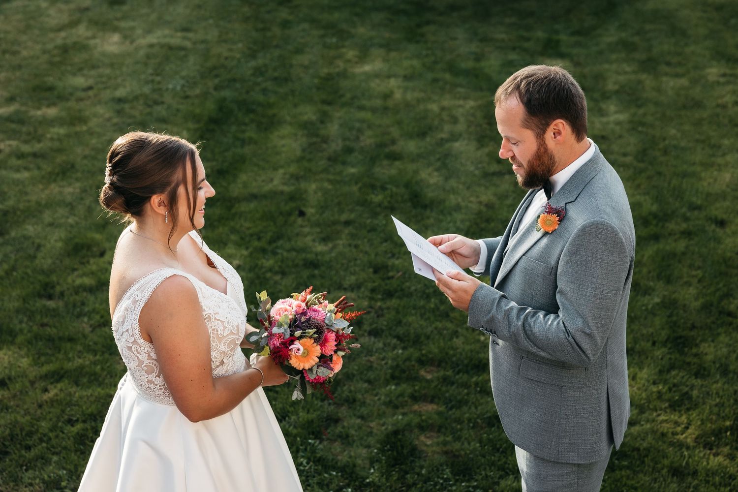 Groom reads heartfelt wedding vows to smiling bride holding colorful bouquet during outdoor private vow exchange ceremony.