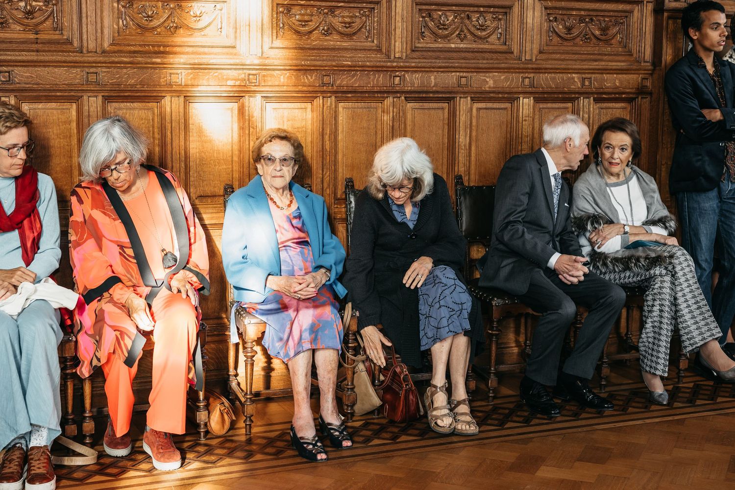 Elderly wedding guests seated on wooden chairs along a paneled wall at a formal ceremony venue.