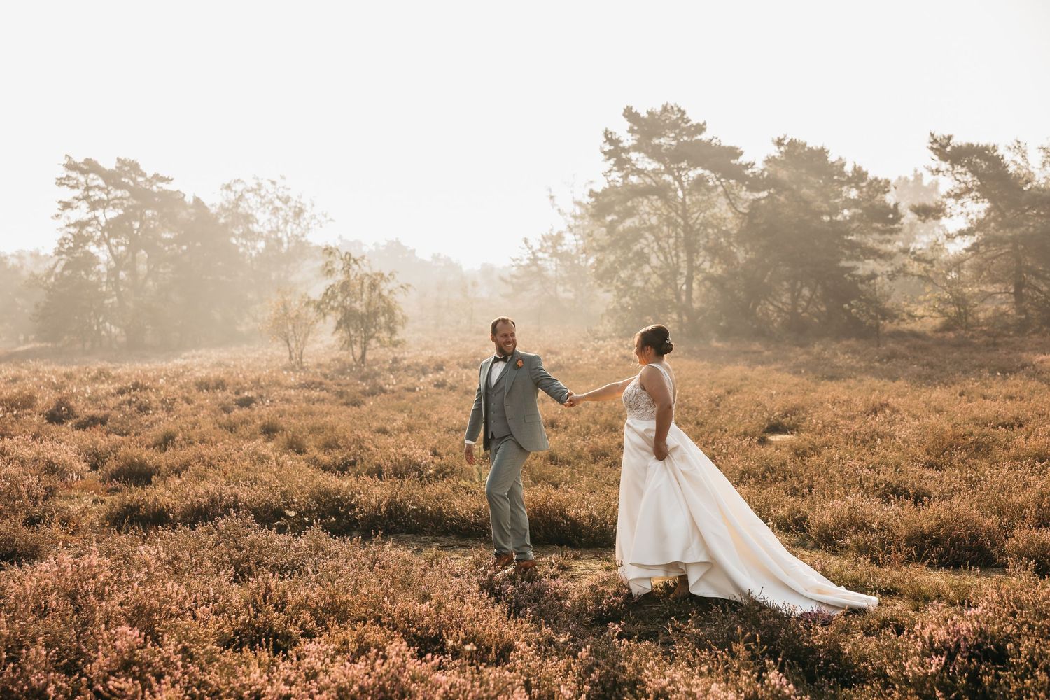 Bride and groom walking hand in hand through sunlit misty heather landscape in wedding attire.