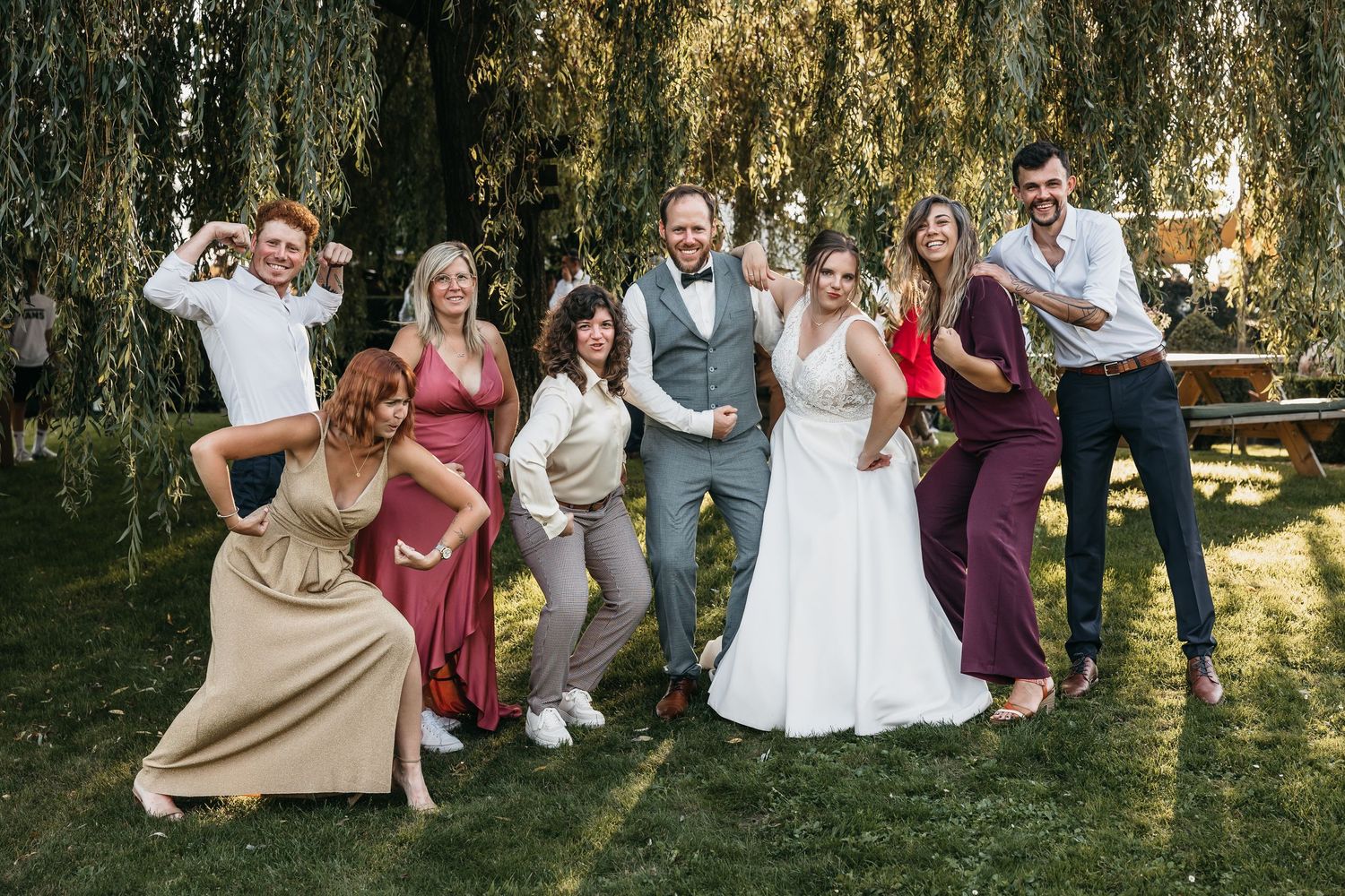 Wedding couple poses playfully with bridal party under weeping willow tree outdoors.