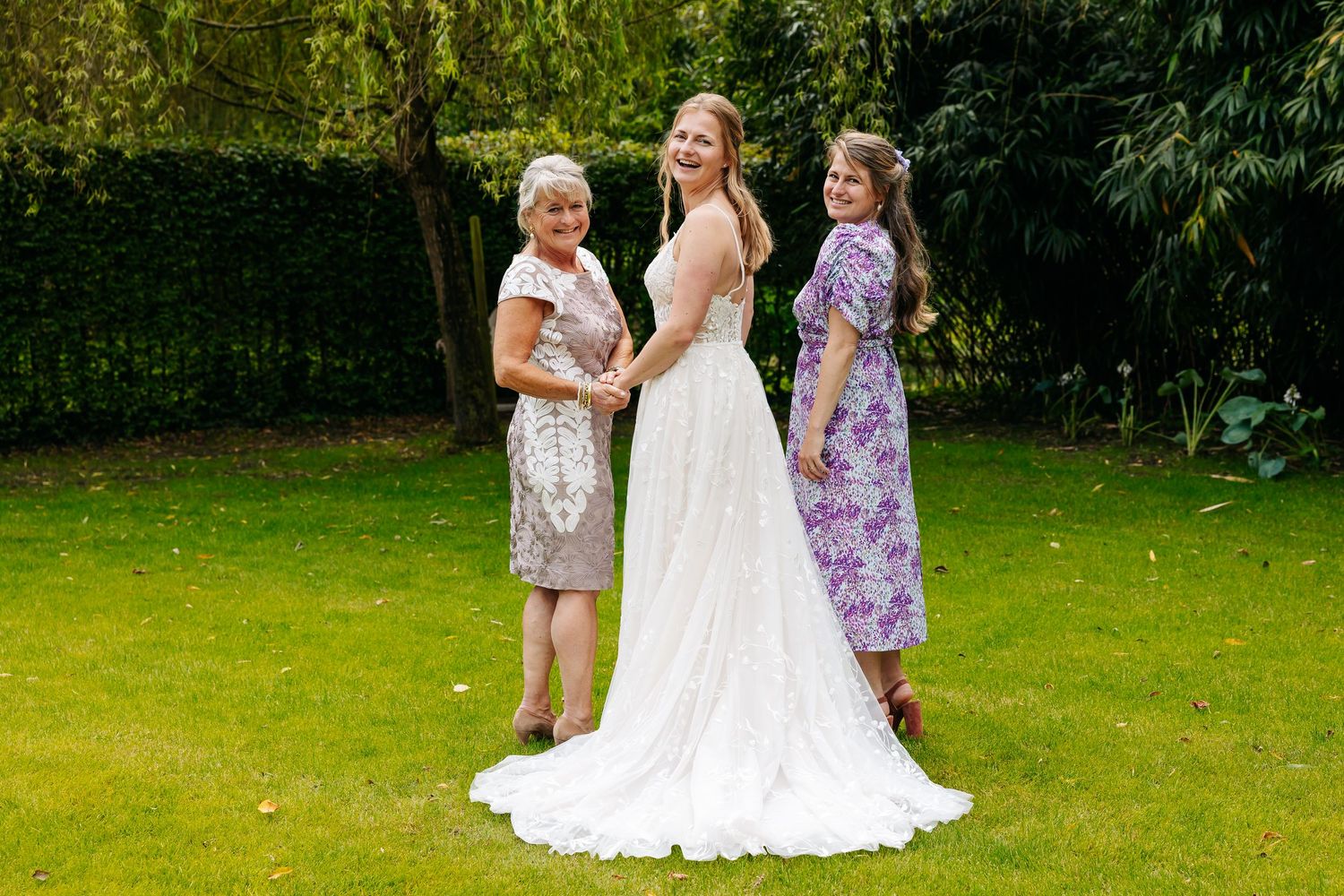 Bride in elegant white gown poses outdoors with two guests in floral dresses in a lush green garden.