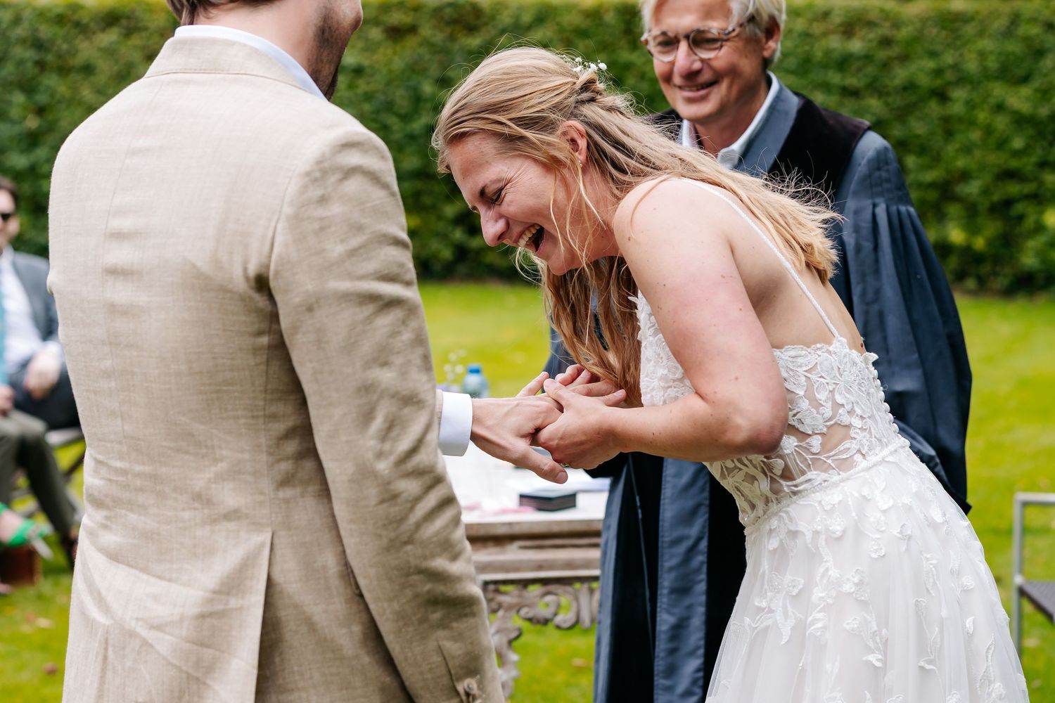 Bride laughing joyfully while exchanging rings with groom during outdoor wedding ceremony.