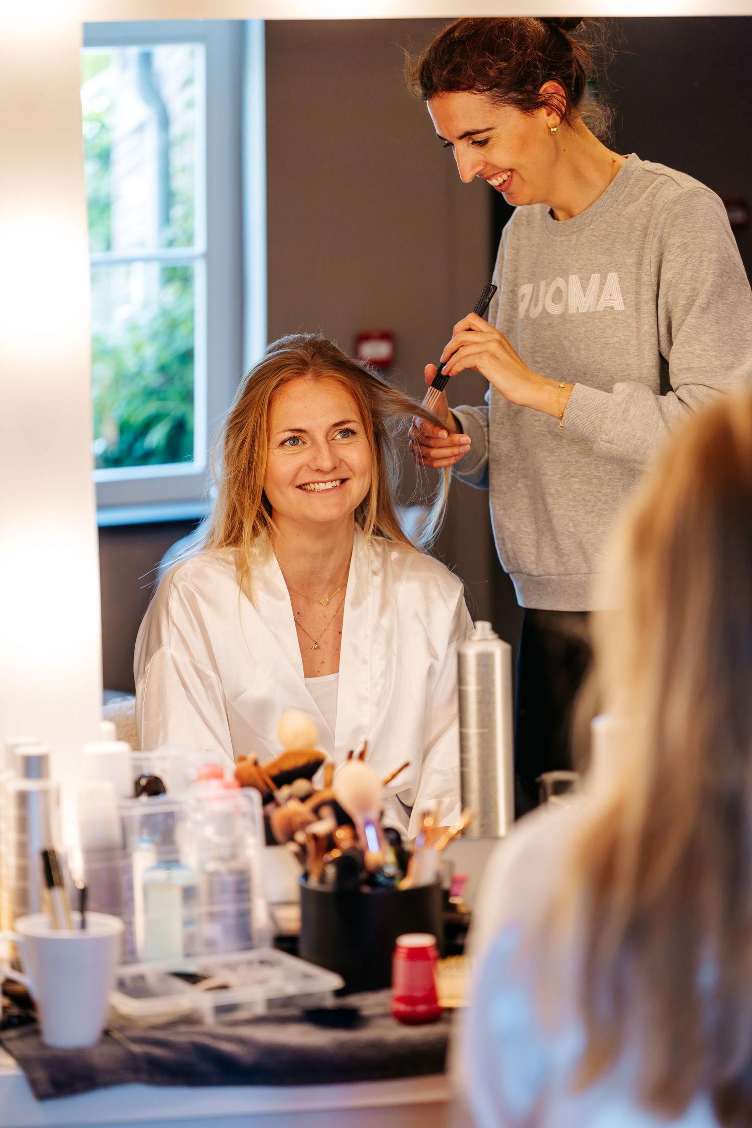 Smiling bride in white robe getting makeup done by artist at vanity mirror with bright lights.