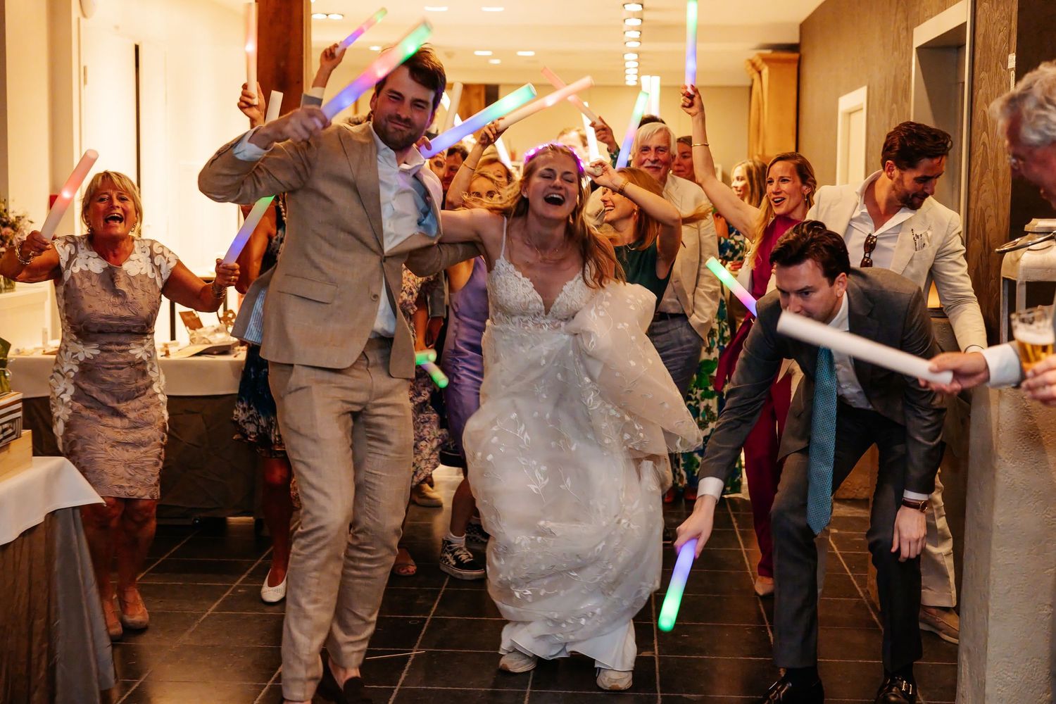 Joyful bride and groom celebrate with guests waving colorful glow sticks at wedding sendoff.