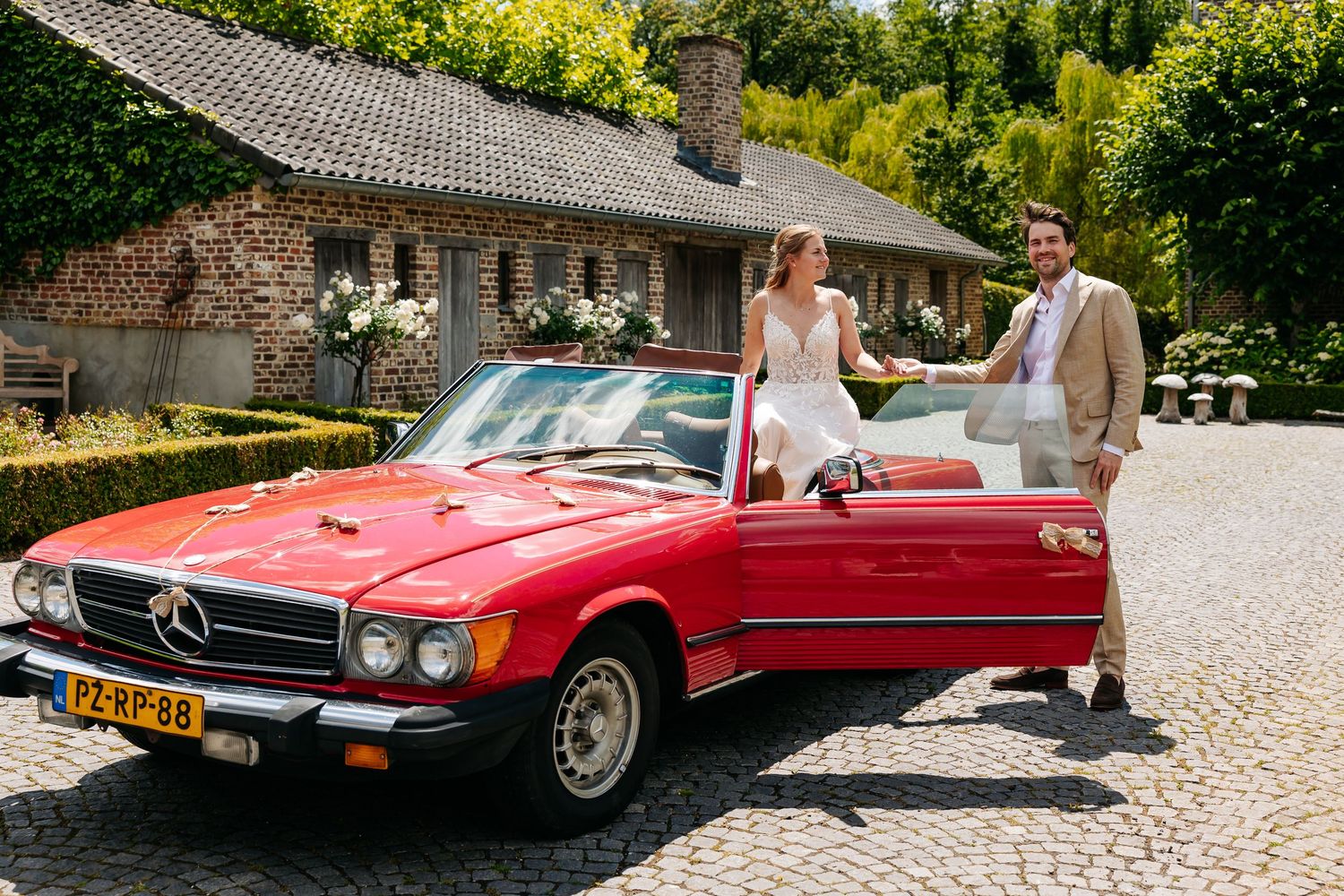 Bride and groom posing beside a classic red Mercedes-Benz convertible on cobblestone drive.
