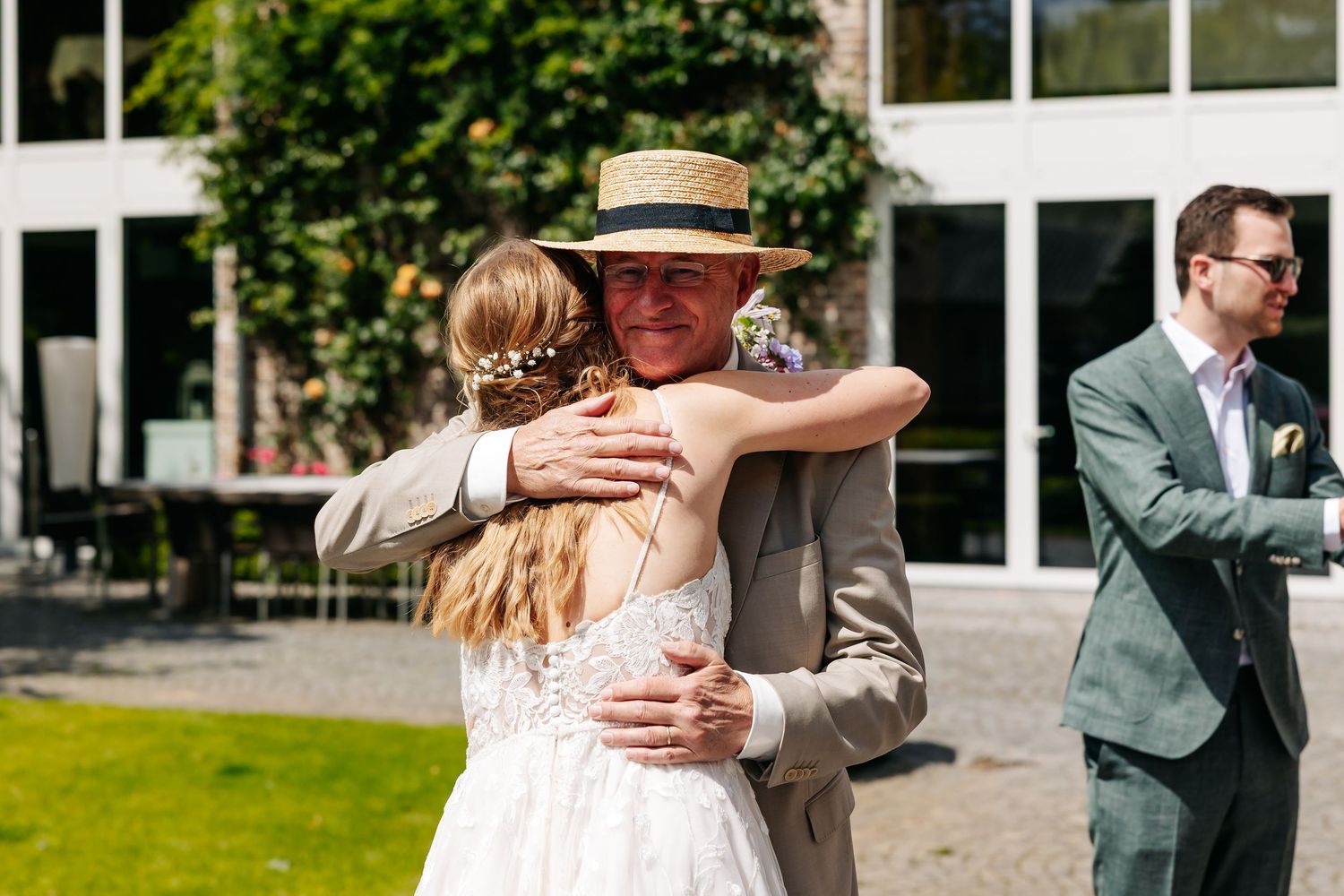 Older man in straw hat warmly embracing bride in white gown at outdoor wedding.