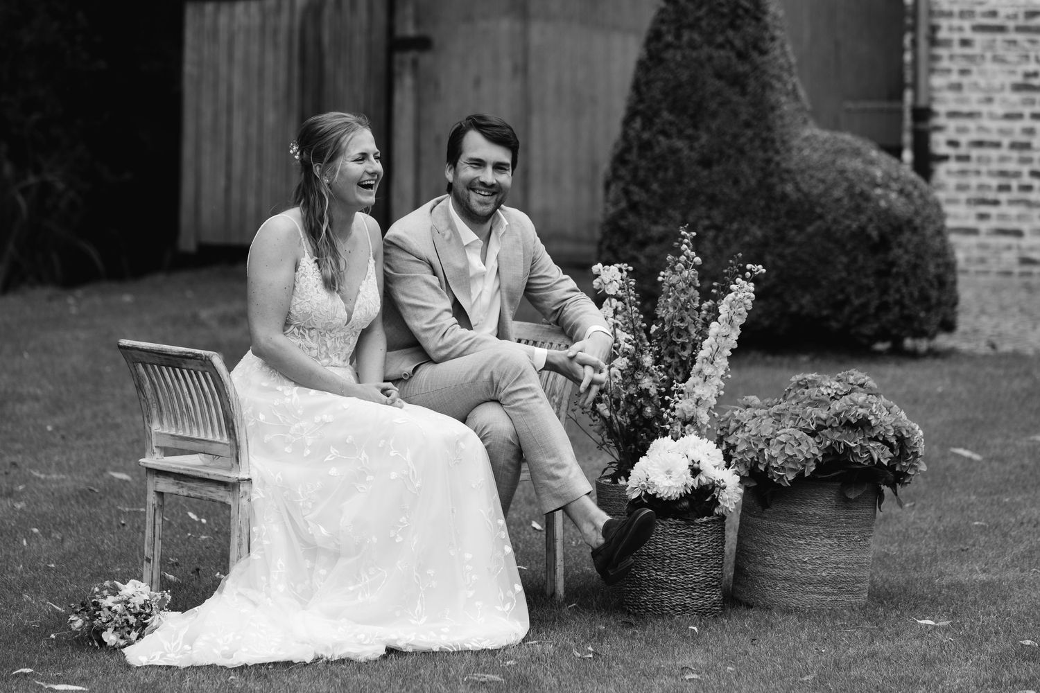 Black and white photo of laughing bride and groom sitting together outdoors with floral arrangement.