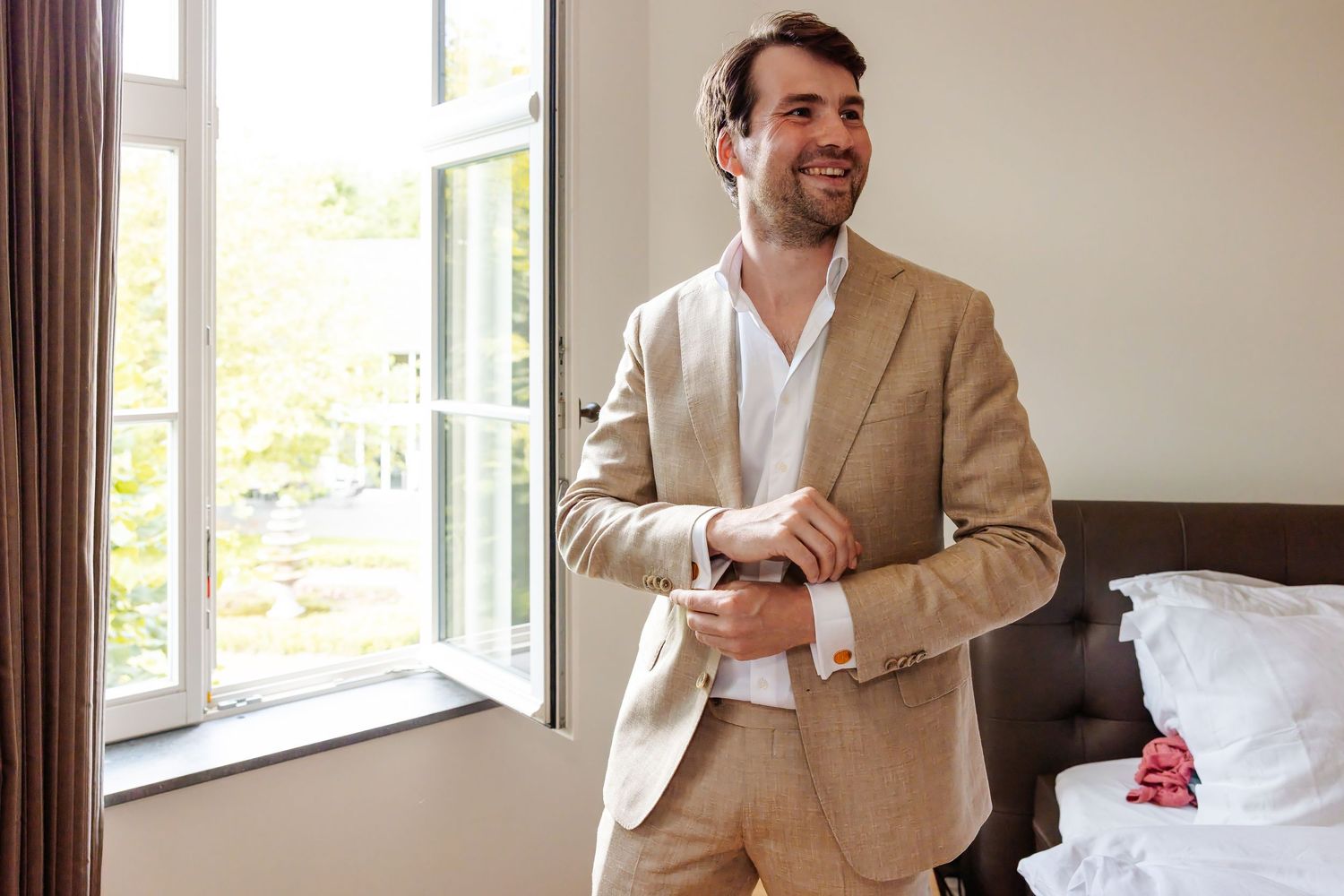 Smiling groom in beige linen suit adjusting cufflinks by an open window in a bright hotel room.