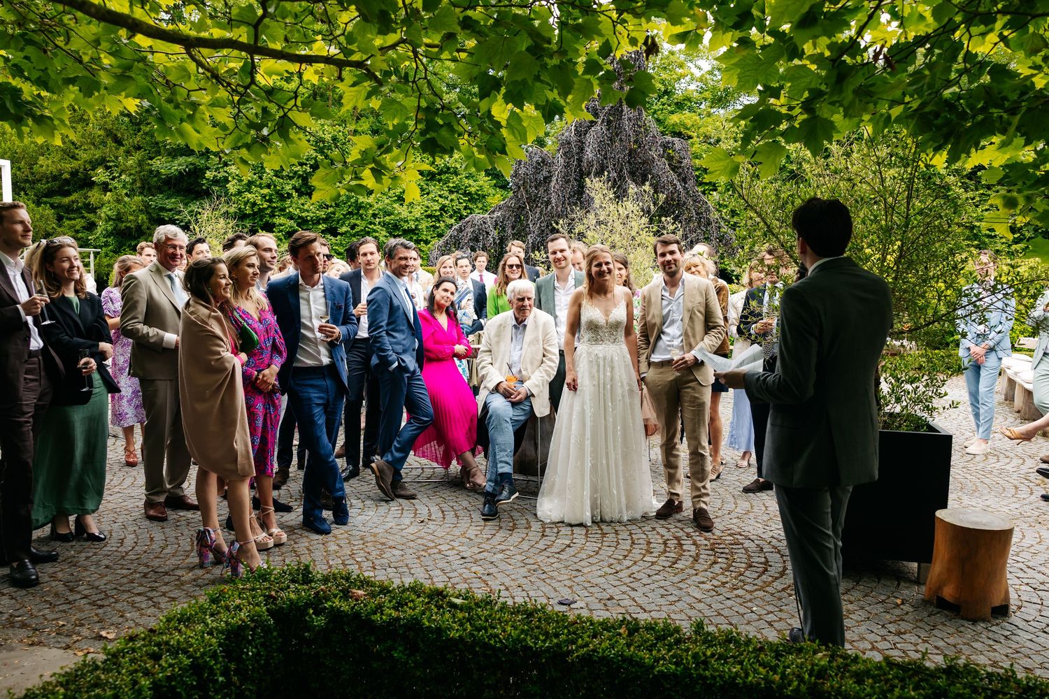 Wedding guests gather outdoors in a green garden to listen to a speech by the groom.