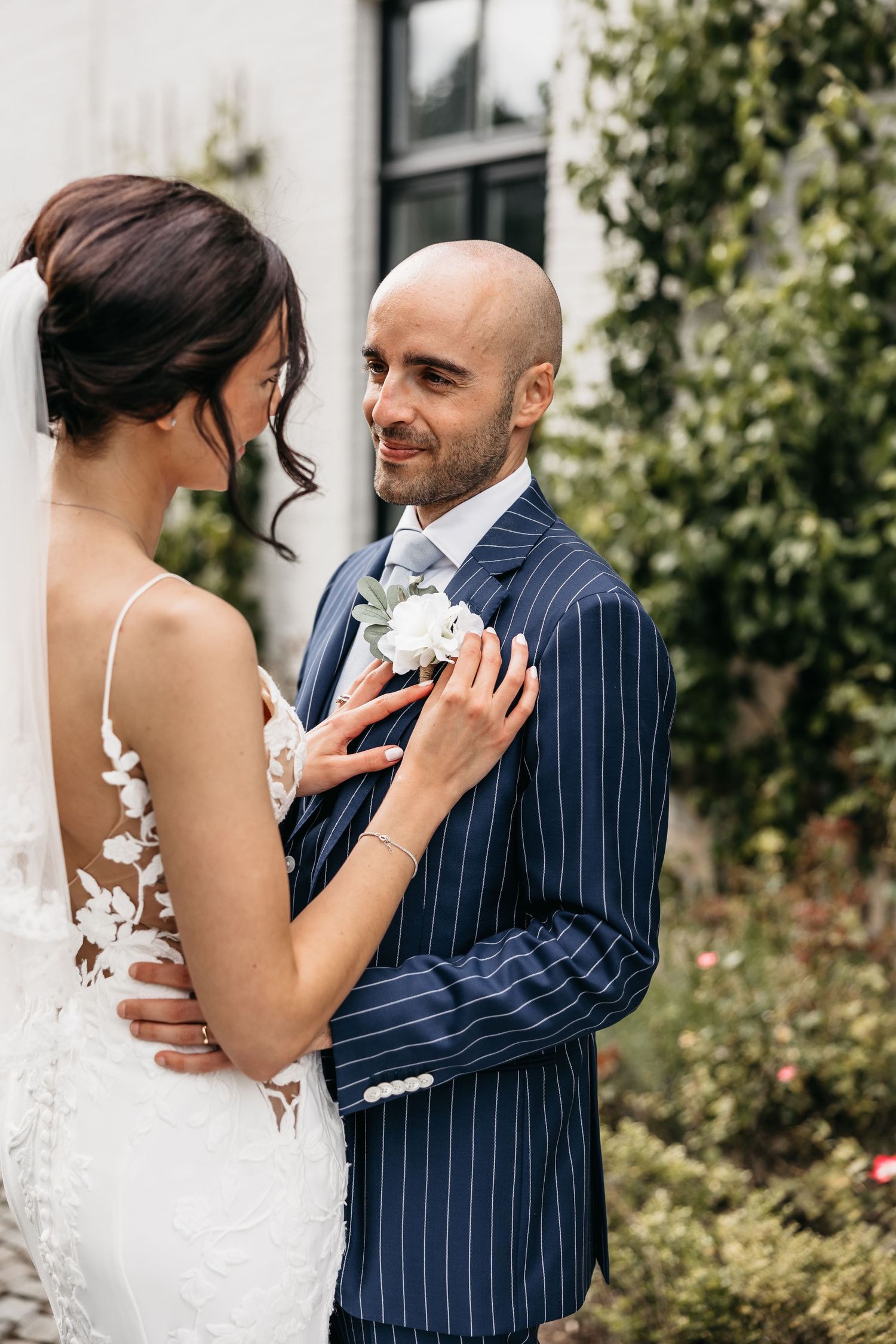 Bride adjusts grooms white boutonniere as he smiles warmly at her in a lush garden setting.