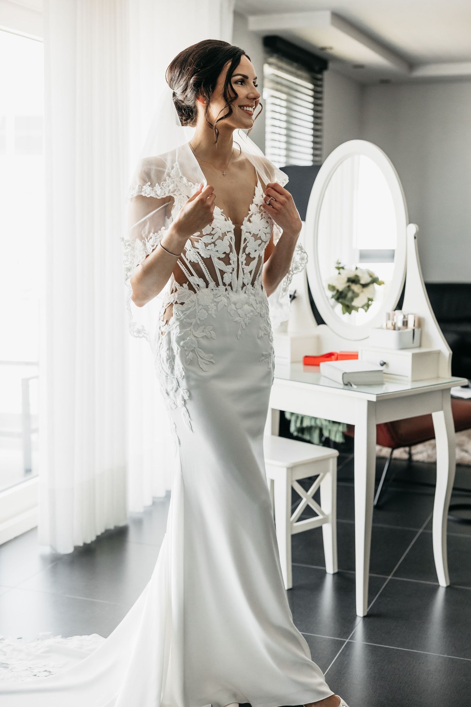 Elegant bride in fitted lace wedding gown smiling beside white oval mirror in bright room.