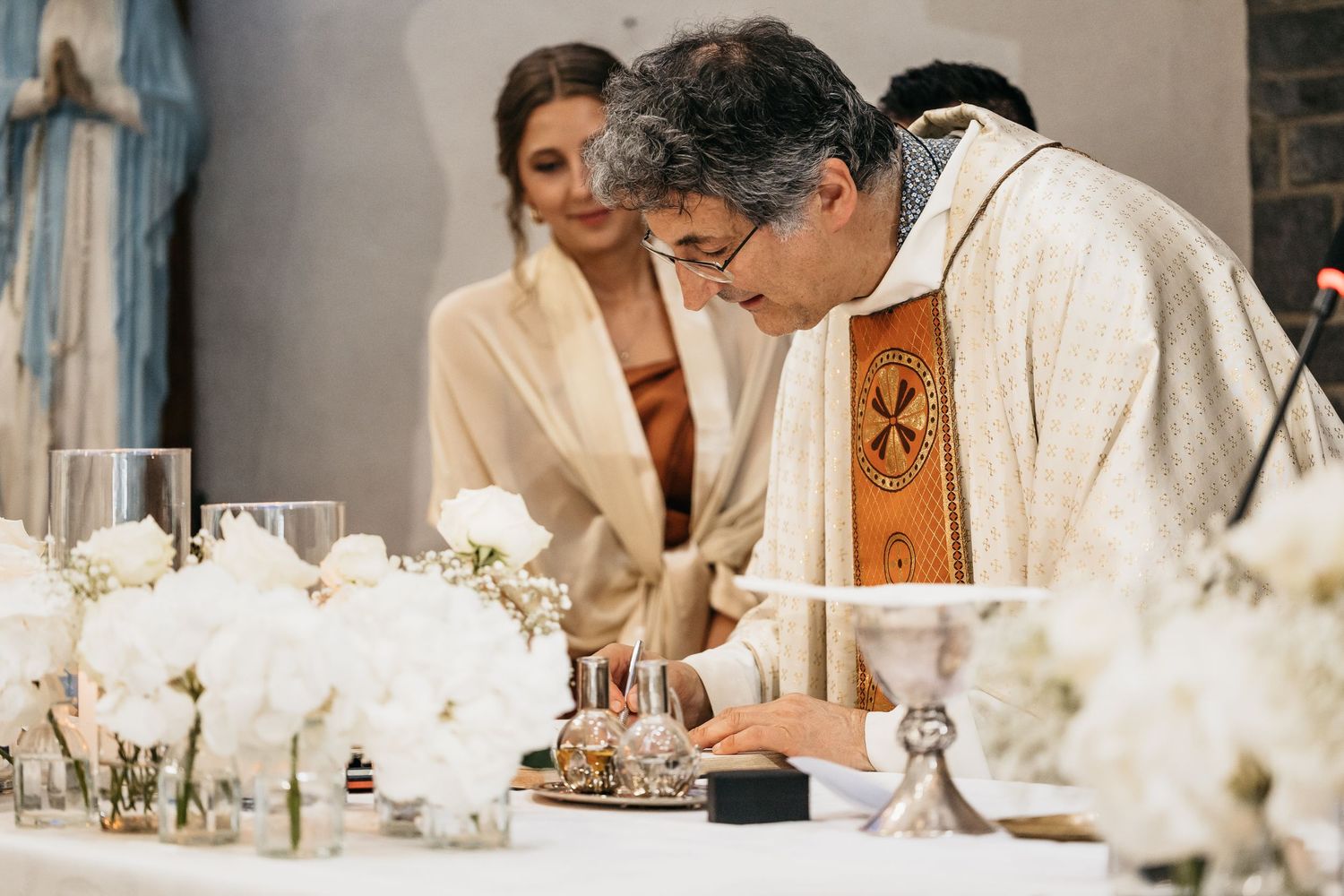 Catholic priest signing wedding documents at white flower-adorned altar during marriage ceremony.