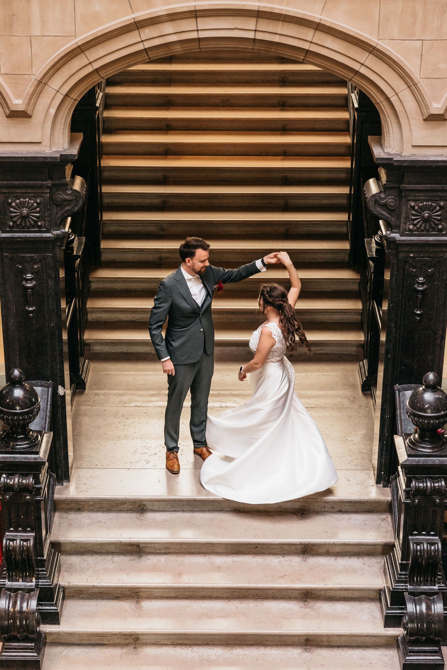 Groom twirling bride in white dress on ornate marble staircase.