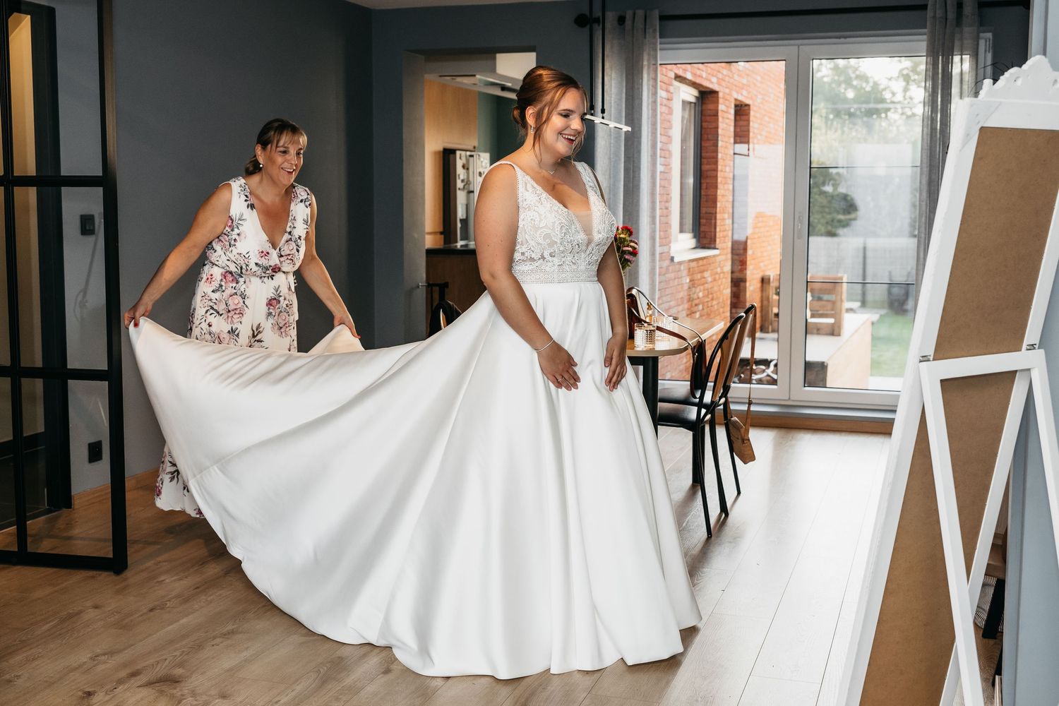 Bride in white A-line wedding gown twirling skirt with helper in floral dress in bright room.