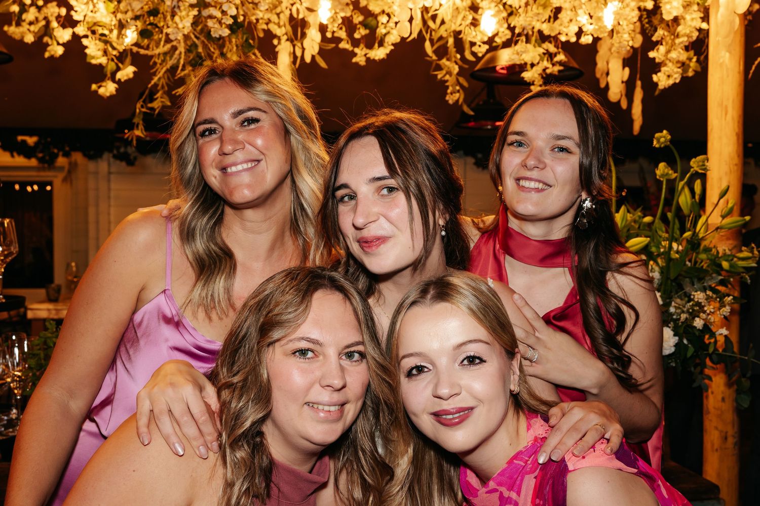 Five young women in pink dresses smiling together under floral decorations at a wedding reception.