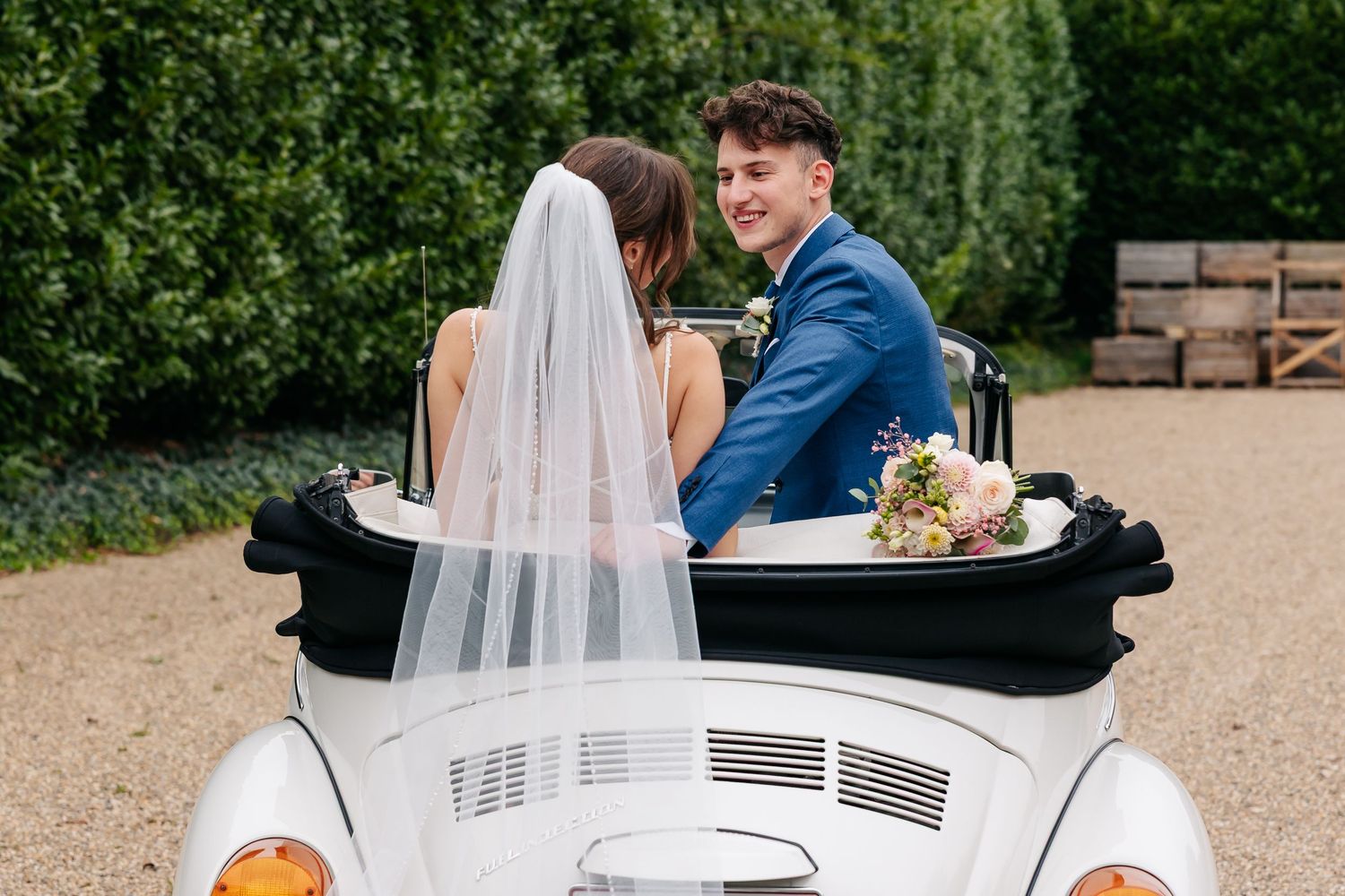 Groom in blue suit smiles at bride holding bouquet while seated in white VW Beetle convertible car.