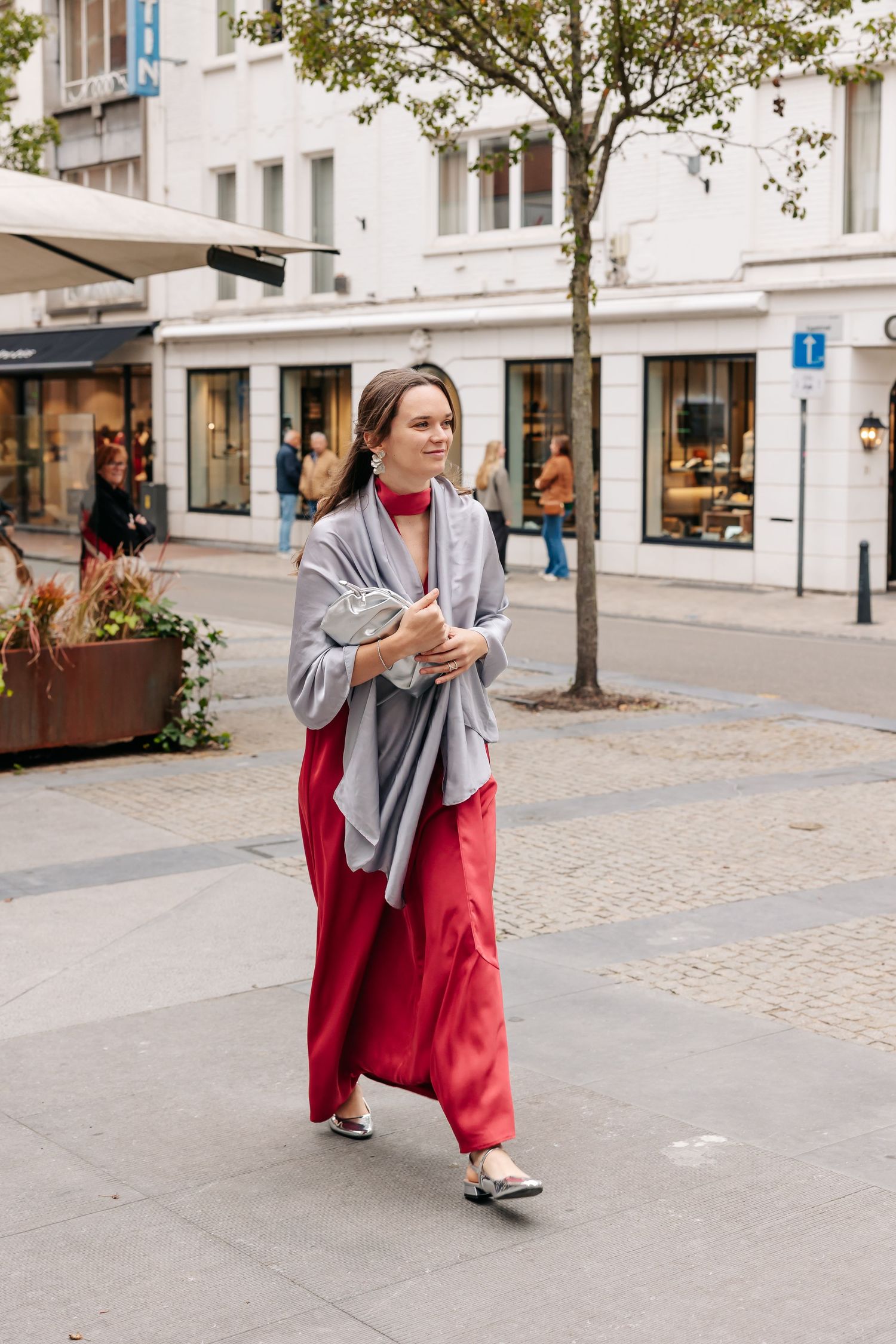 Woman in red wide-leg pants and gray shawl walking along a European city street carrying a clutch purse.