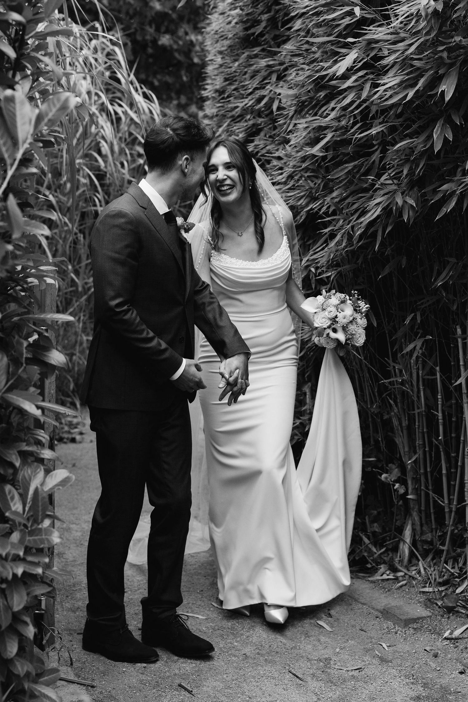 Black and white photo of smiling bride and groom walking hand in hand through lush garden path.