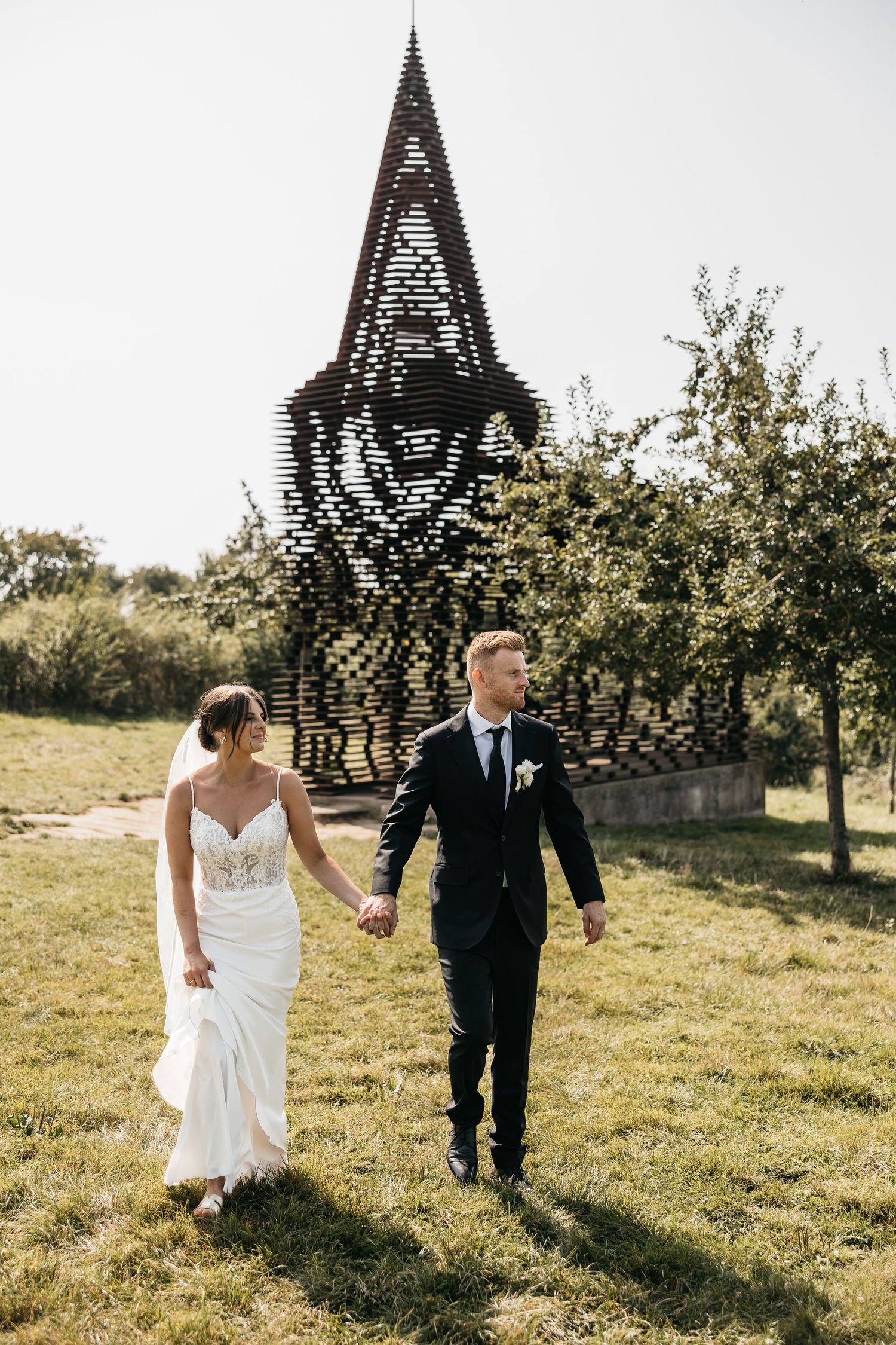 Bride and groom walking hand in hand on green grass with a latticed church tower in background.