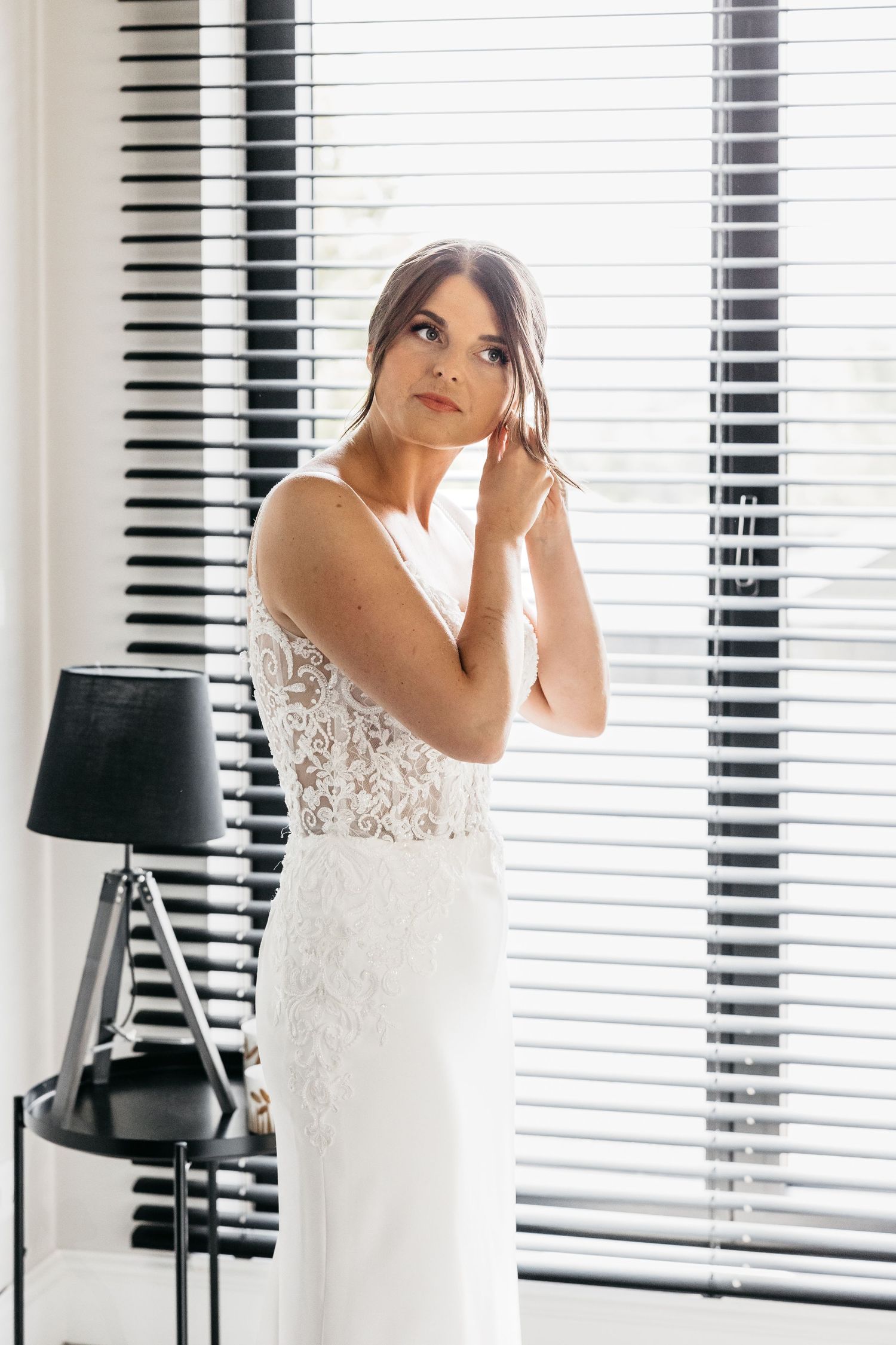 Bride in white lace wedding dress standing by window with venetian blinds adjusting her hair.