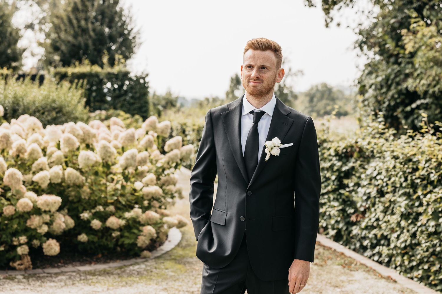 Stylish groom in black suit with white boutonniere posing beside blooming hydrangeas.