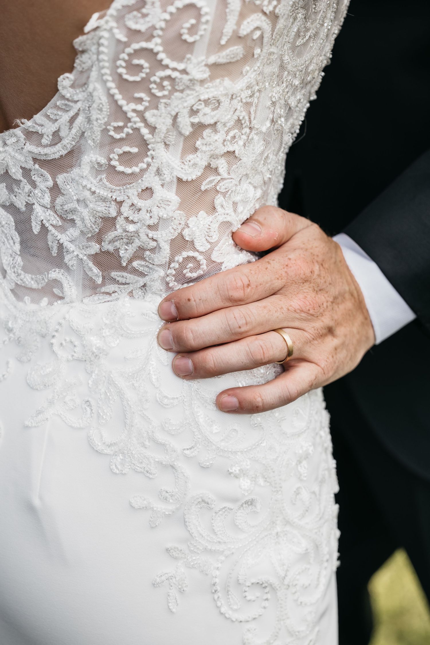 Groom's hand with gold wedding band resting on bride's white lace wedding dress.