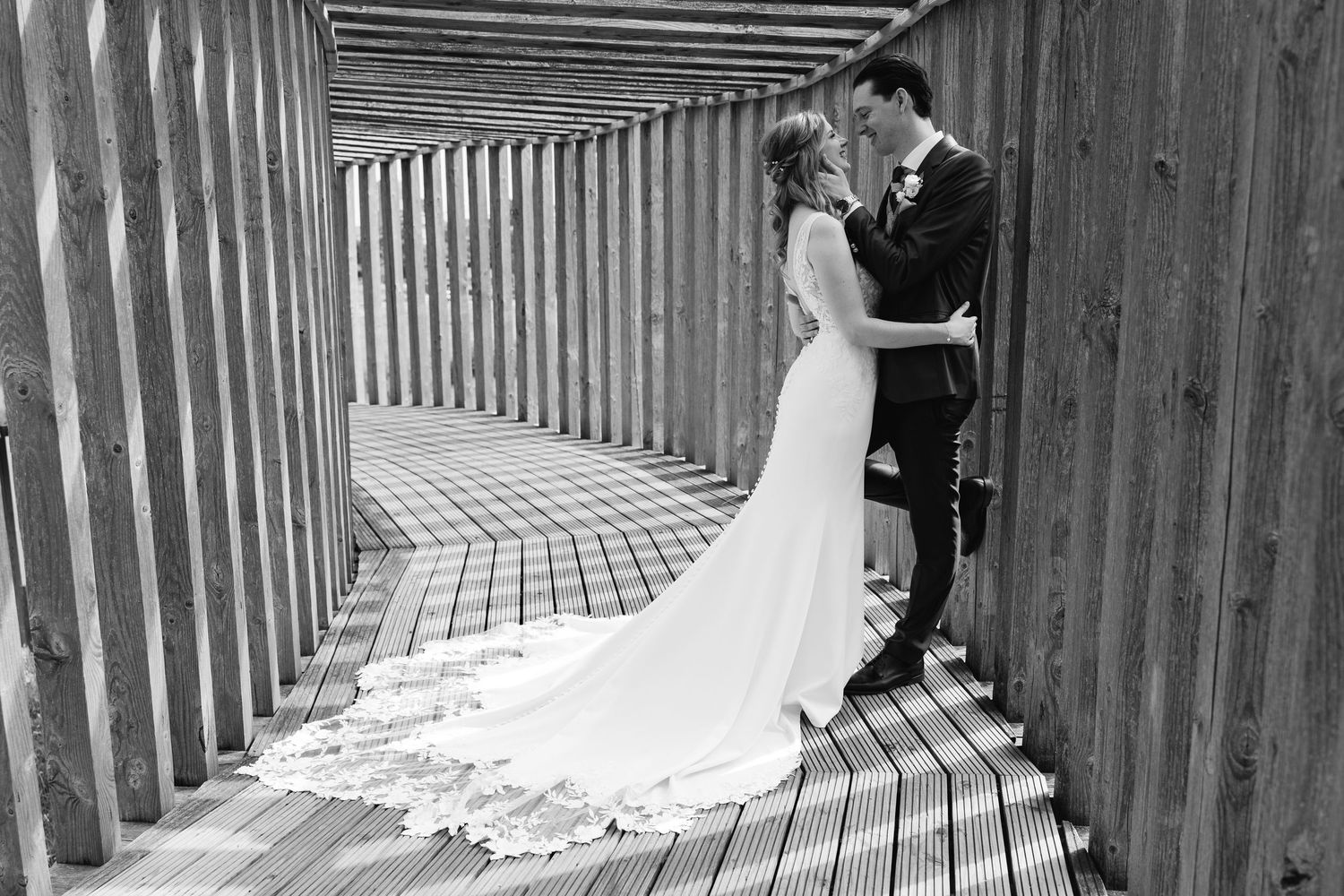 Black and white photo of bride and groom embracing in wooden pergola walkway.