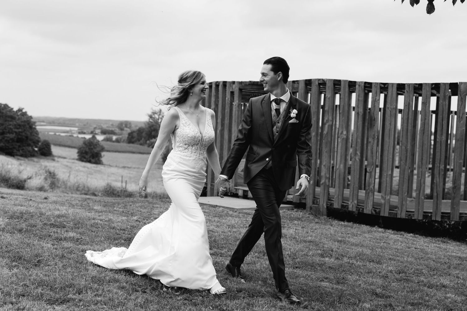 Black and white photo of a bride and groom walking hand in hand along a grassy hillside on their wedding day.