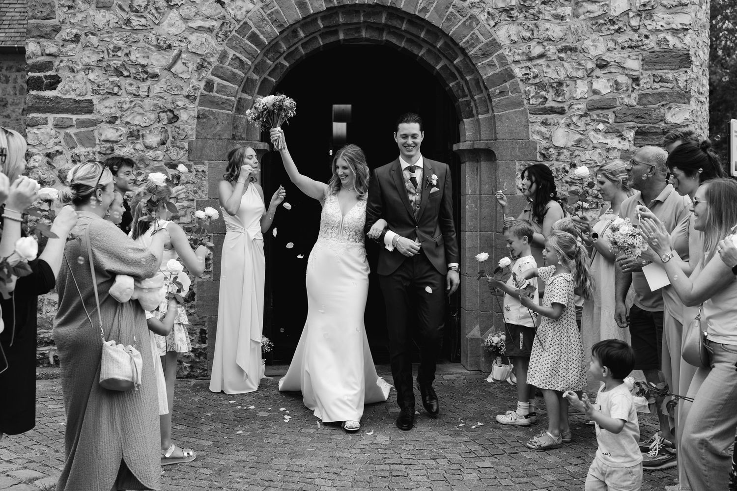 Black and white image of newlyweds walking through stone church arch with celebrating guests.