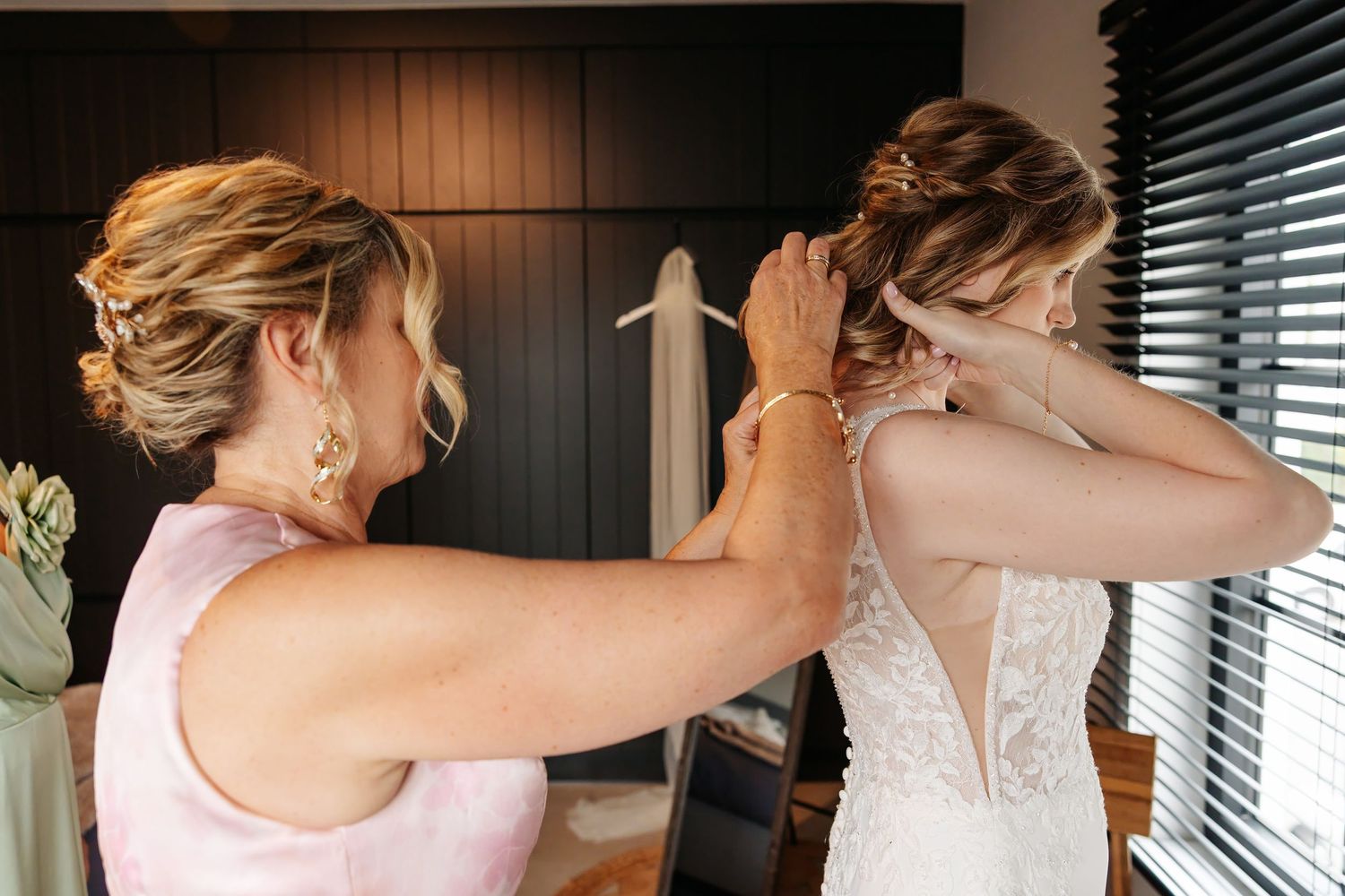 Bridesmaid in pink dress fastening a delicate pearl necklace on a bride in a backless lace wedding gown.
