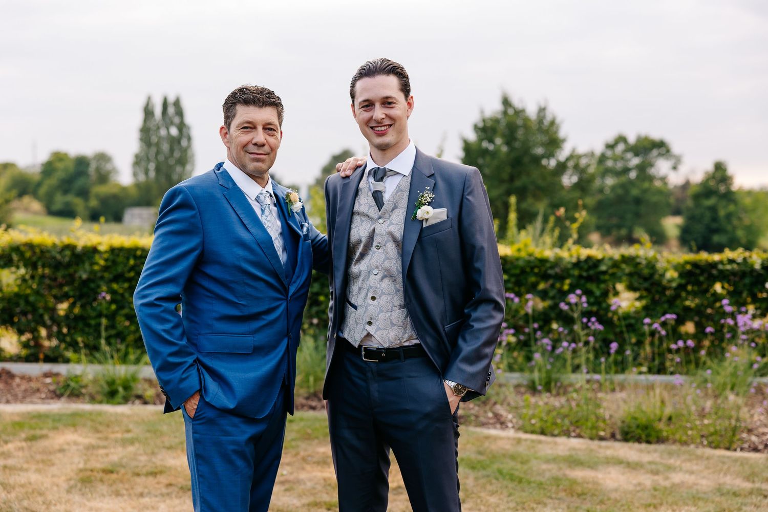 Two men in suits smile outdoors at a wedding, one in blue and one in navy with a floral boutonniere.