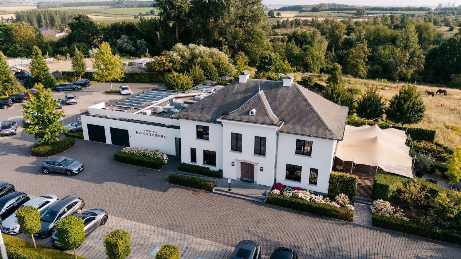 Aerial view of elegant white wedding venue building with parking lot surrounded by green countryside.