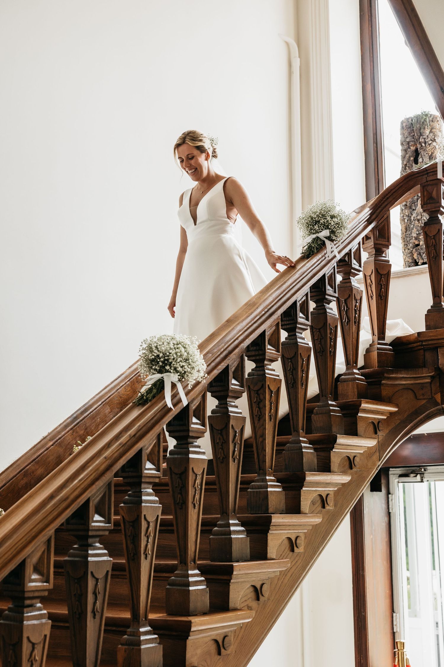 Elegant bride in white gown descending ornate wooden staircase decorated with floral arrangements.