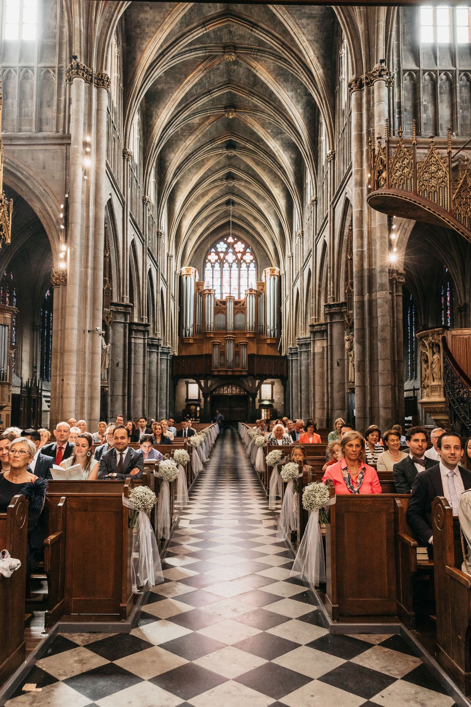 Full cathedral interior view of wedding ceremony with guests seated in wooden pews beneath Gothic vaulted ceiling.