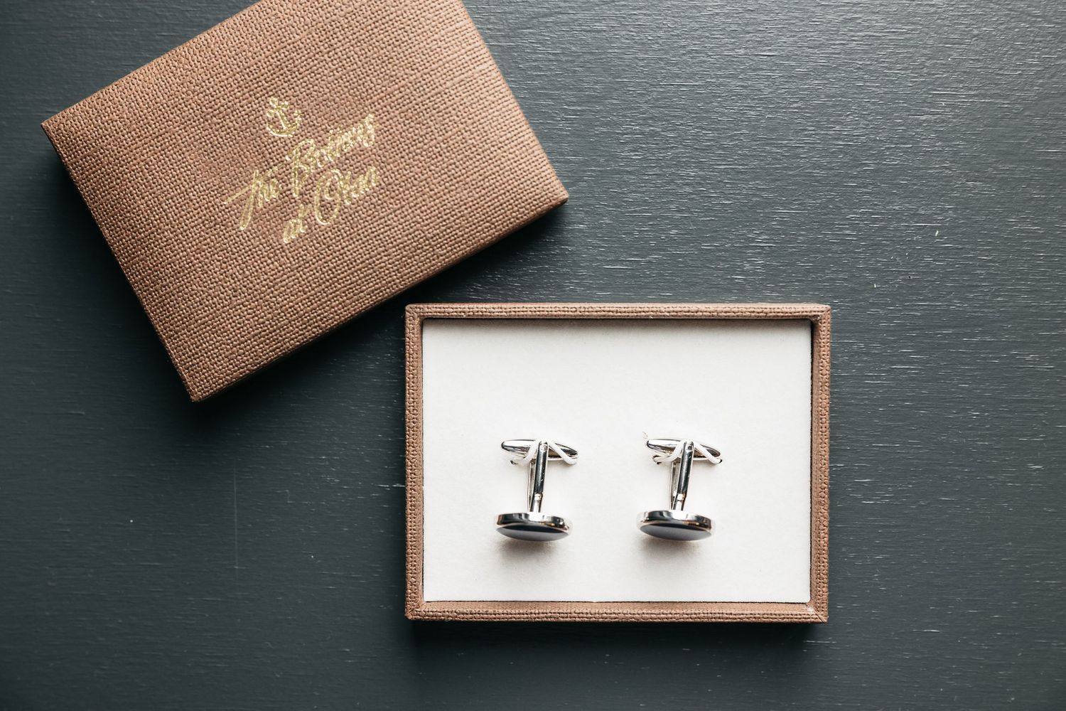Silver cufflinks displayed in a brown gift box with gold text on a dark surface.